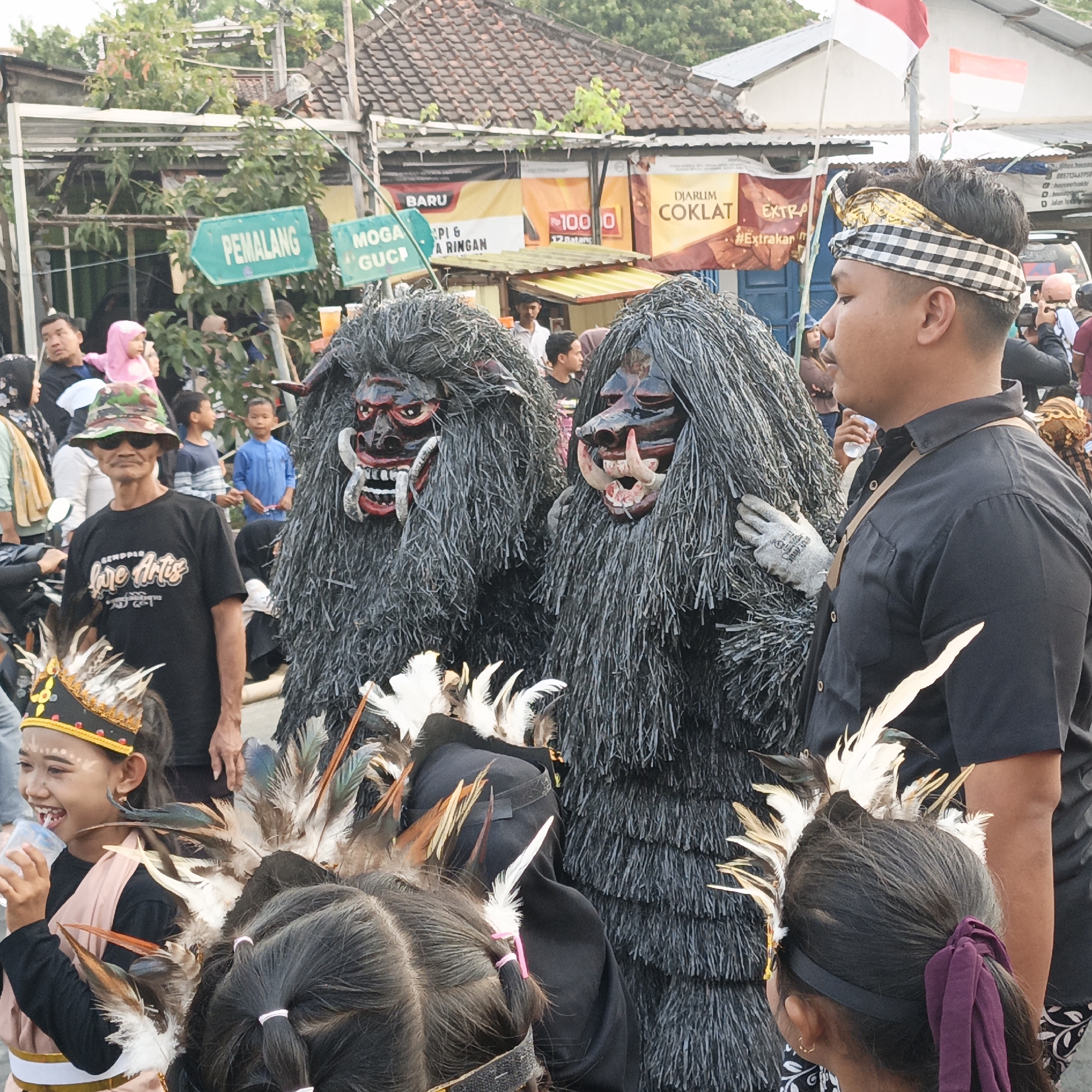 In Randudongkal, Indonesia, a street scene unfolds with a procession or performance. Two individuals, adorned in tall, shaggy grey costumes with intimidating red-and-black masks resembling monstrous faces with bared teeth and fangs, are the central focus. One mask has a more open mouth, revealing a row of white teeth. Their bodies are almost entirely concealed by the textured material.
To the right, a young man in a black short-sleeved shirt, wearing a black and white checkered headband, stands in profile. His face is turned to the side, and he appears to be observing the figures. He has a light brown strap across his chest. He is wearing a grey glove on his left hand which is resting on the costume to his left.
In the foreground, a young girl with a cheerful expression is being held or positioned. She wears a black shirt beneath a pale pink sleeveless garment adorned with a golden belt and trim. On her head, she sports a small headdress with colorful feathers and what appears to be gold trim. She is holding a transparent cup with a white lid to her mouth. Behind her, other children are visible, including one with a more elaborate headdress of feathers and dark hair tied with pink bands.
A man in a camouflage patterned hat and sunglasses, wearing a black t-shirt with white text, stands to the left, looking towards the girl. Several other people, including adults and children, are visible in the background, some blurred and out of focus.
Signs are posted in the background, including a green sign with "PEMALANG" in white text, indicating a location or direction. Other banners and structures suggest a marketplace or event setting. The ground appears to be paved. The overall atmosphere seems to be one of a cultural event or festival.