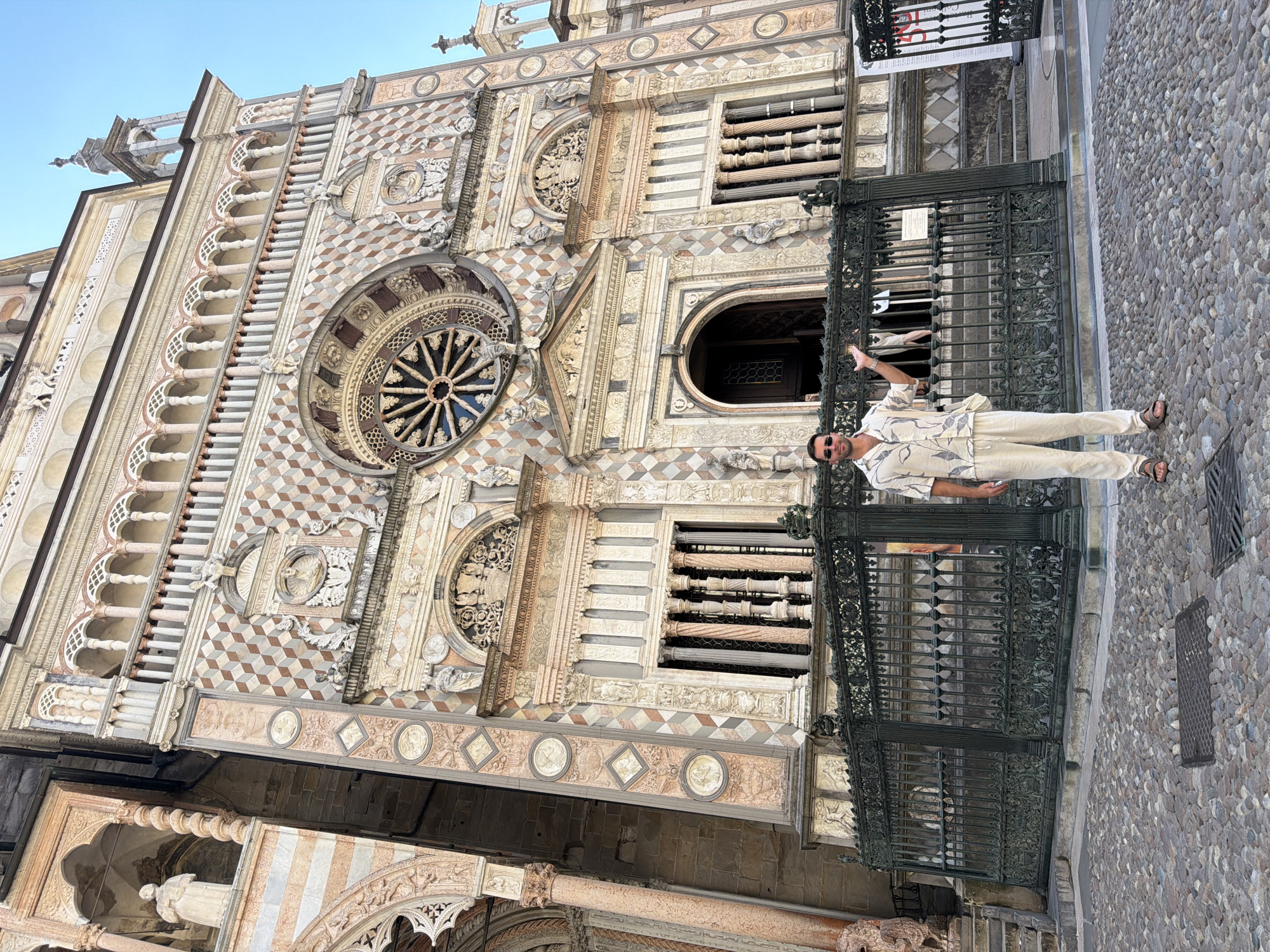 A person stands on a cobblestone street in front of a large, ornate building with intricate architectural details. The building, likely a cathedral or similar historical structure, features a rose window, arched doorways, and decorative stonework. The person is wearing a light-colored patterned shirt and pants, sunglasses, and sandals. They are gesturing with their left hand, pointing towards the building. A black wrought-iron gate separates the street from the building's entrance. The sky above is clear and blue, suggesting a bright, sunny day. The time of day appears to be late morning or early afternoon. The location is identified as Mozzo, Italy. No visible text is present in the image.