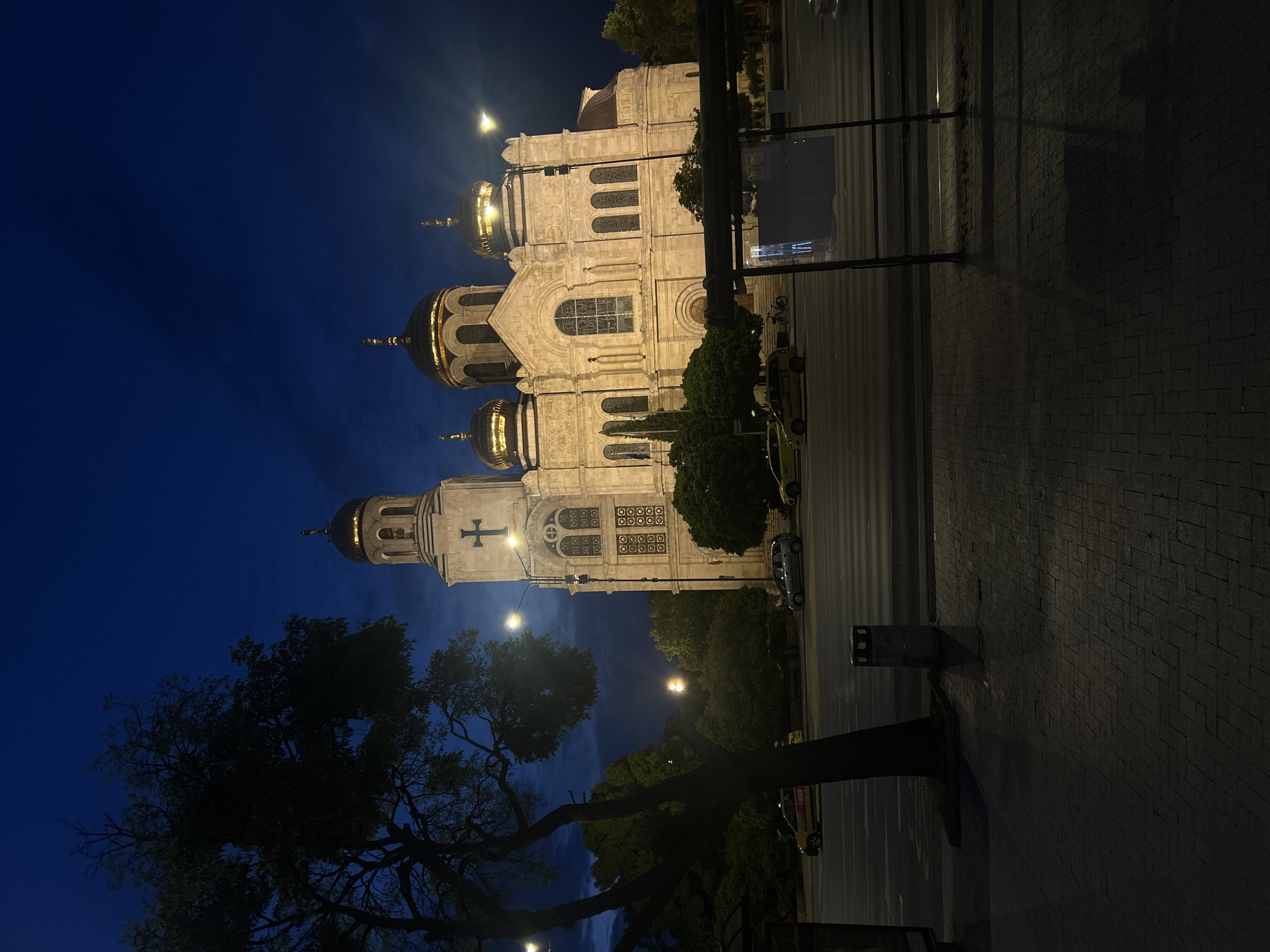 The image captures a grand, illuminated church at night, set against a deep blue sky in Varna, Bulgaria. The central subject is a large Orthodox cathedral, constructed from light-colored stone, featuring multiple prominent golden domes and crosses, which reflect the artificial lighting. The church's facade displays intricate architectural details, including arched windows with decorative grilles and pilasters. One section of the church, possibly a bell tower, clearly shows a cross at its peak.
In the foreground and to the left of the church, the silhouettes of large, dark trees with visible branches and some foliage create a stark contrast against the lit building and night sky. Streetlights cast bright glows, illuminating parts of the scene and creating lens flares.
A paved street or road runs in front of the church, where several vehicles, including at least two yellow taxis, are visible. A bicycle is also parked near the church on what appears to be a sidewalk or paved area. Street furniture, such as a dark bollard and a tall, dark rectangular sign or billboard, are present on the right side of the scene. The text on the billboard is unreadable due to distance and lighting.
The overall atmosphere is serene and majestic, highlighting the architectural beauty of the church under the night sky. The scene depicts a typical urban nighttime view, emphasizing the landmark's presence.