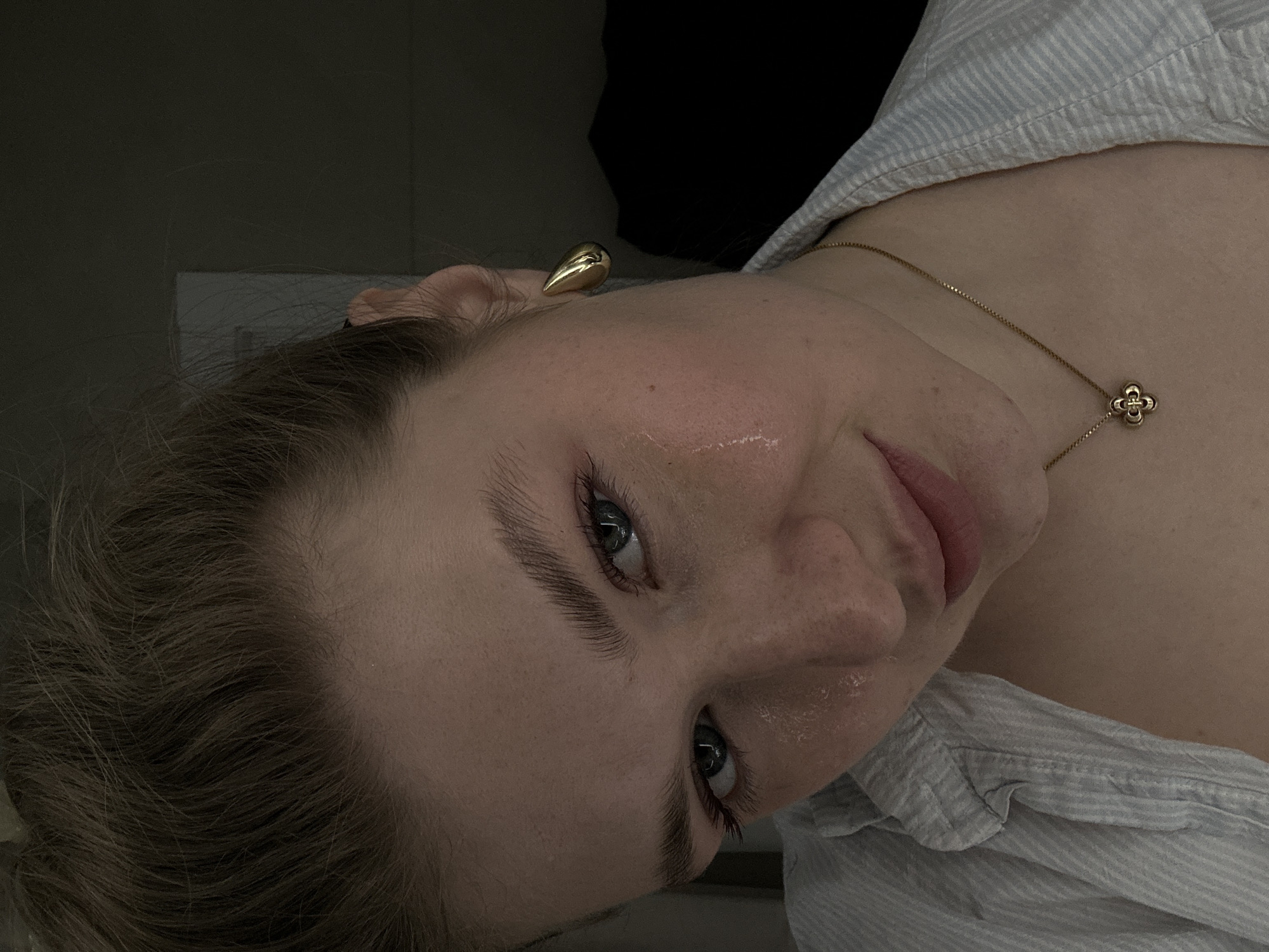 A close-up, rotated image depicts a person with fair skin, brown hair, and blue eyes. They are wearing a light blue striped shirt and a gold necklace with a floral pendant. A gold teardrop earring is visible in their left ear. The background includes a dark, indistinct area and a section of a white wall. The location is not discernible from the image.