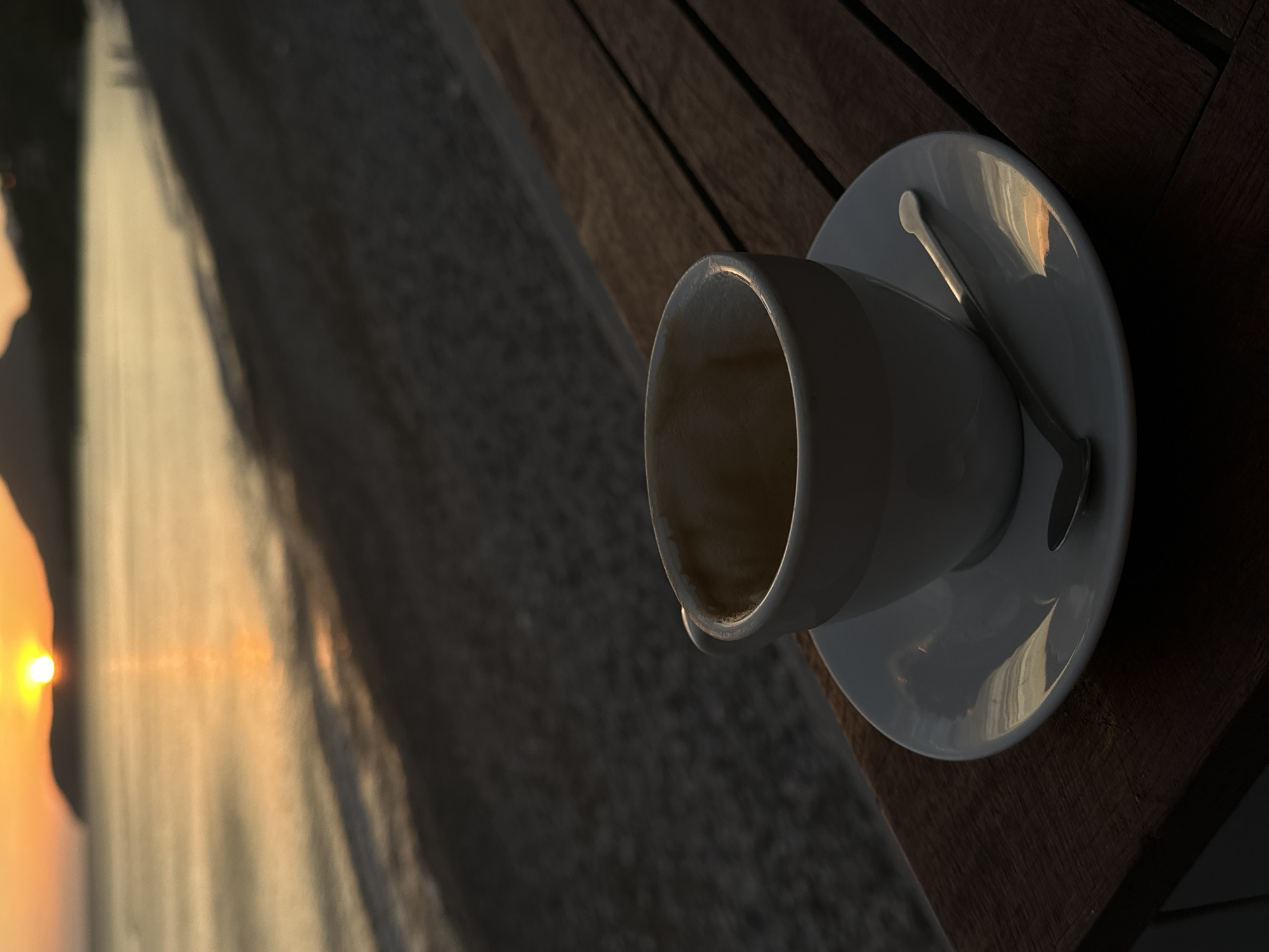 The image depicts an empty coffee cup and saucer with a spoon on a dark wooden table, likely outdoors. In the background, a blurred image of what appears to be a shoreline at sunset is visible. The sun's reflection is prominent in the water, and the overall scene suggests a location near a beach. Given the reference to Bar, Montenegro, it is highly probable the photograph was taken at a cafe or restaurant overlooking the Adriatic Sea in or near Bar. There are no people visible in the immediate scene. The focus is on the remnants of a beverage and the adjacent landscape, suggesting a recent activity of enjoying a coffee with a view.