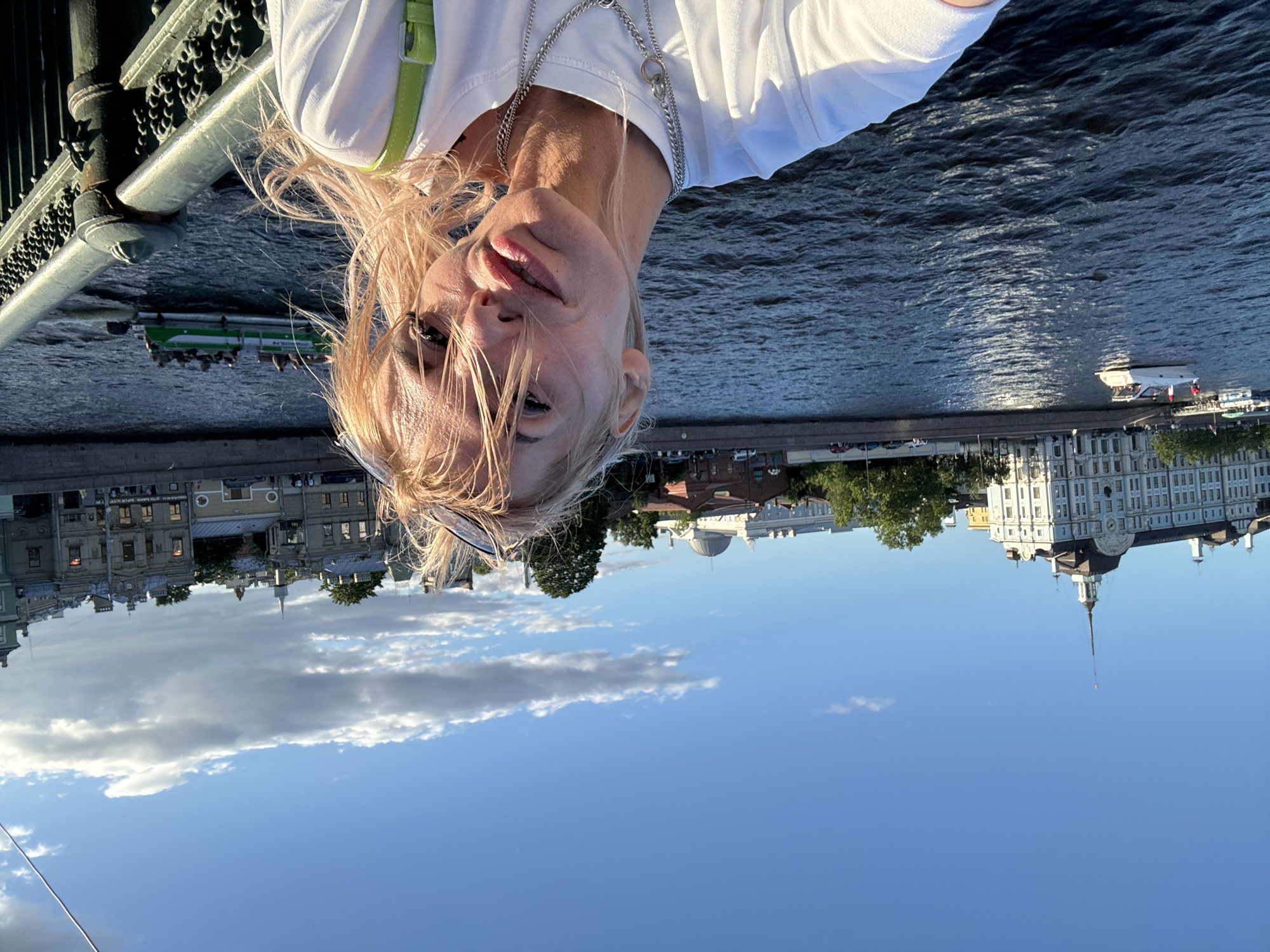 A woman is taking a selfie on a bridge over a wide river in St. Petersburg, Russia. She has blonde hair, is wearing a white t-shirt and a green strap is visible over her shoulder. In the background, there are several boats on the water and buildings along the embankment. The sky is blue with some clouds. The railing of the bridge is visible in the foreground.