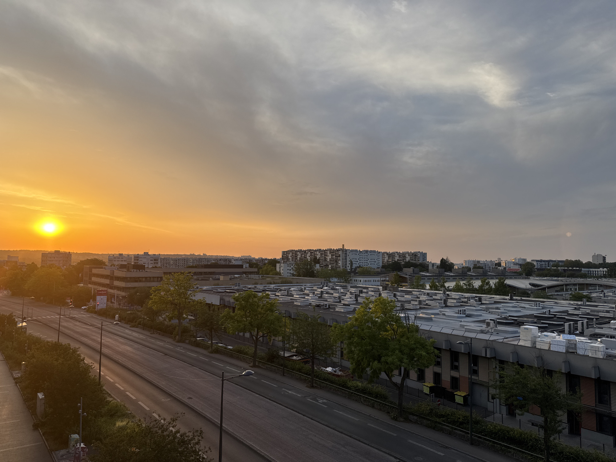 A wide shot captures an urban landscape at dusk. The setting sun, a vibrant orange orb, dominates the left side of the frame, casting a warm glow across the sky. The sky transitions from a bright orange near the horizon to a muted grey and blue at the top. A multi-lane road runs diagonally through the scene, bordered by trees and buildings. In the foreground, the road appears to be empty, with only streetlights and some parked cars visible. Beyond the road, a large industrial or commercial building with a flat roof, adorned with numerous air conditioning units, stretches across the middle ground. Further in the distance, a dense cluster of apartment buildings forms the city's skyline, with a hazy, pale moon visible in the upper right quadrant of the sky. The overall impression is of a quiet evening in a developed urban area.