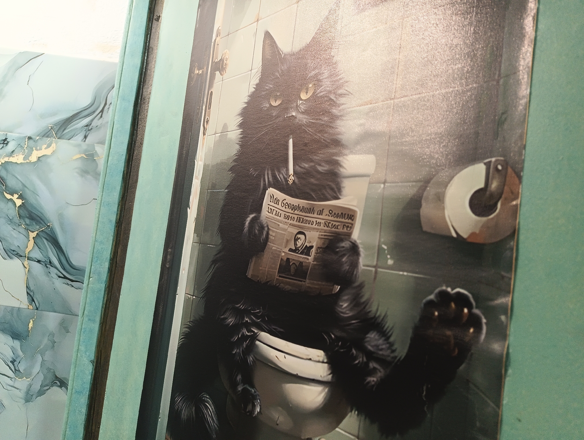 The image is a close-up of a painting depicting a fluffy black cat sitting on a toilet and smoking a cigarette while reading a newspaper. The cat is positioned centrally in the frame, with its tail wrapped around the toilet seat. The newspaper is held in the cat's paws, and its gaze is directed towards the paper. Behind the cat, the wall appears to be tiled with light green tiles, and a roll of toilet paper is visible to the right. The overall scene is enclosed within a frame. The provided context places this painting in the city of Karnobat, Bulgaria, suggesting it is a piece of art displayed in a private or public space within that location.