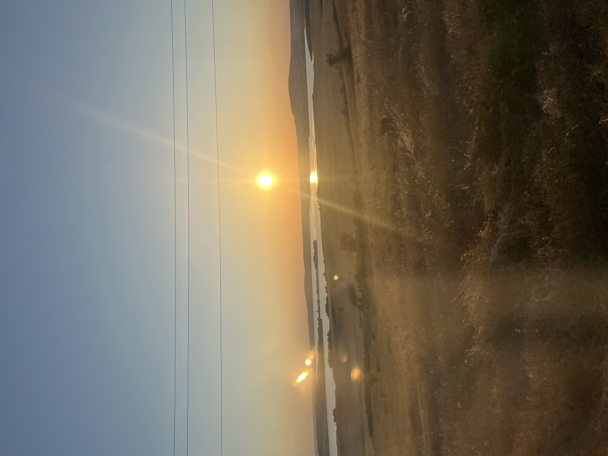 The image depicts a landscape scene during sunset in Tvarditsa, Bulgaria. The sun is low on the horizon, casting a warm, golden light across the sky and reflecting on a body of water that winds through the landscape. The terrain is characterized by rolling hills covered in dry, golden grass. A few trees are sparsely scattered across the fields, and a power line with three parallel wires runs across the left side of the frame, extending into the distance. The sky transitions from a clear blue on the left to a hazy, orange glow near the sun. The overall atmosphere is serene and tranquil, capturing the beauty of a rural sunset. There is no visible text in the image.