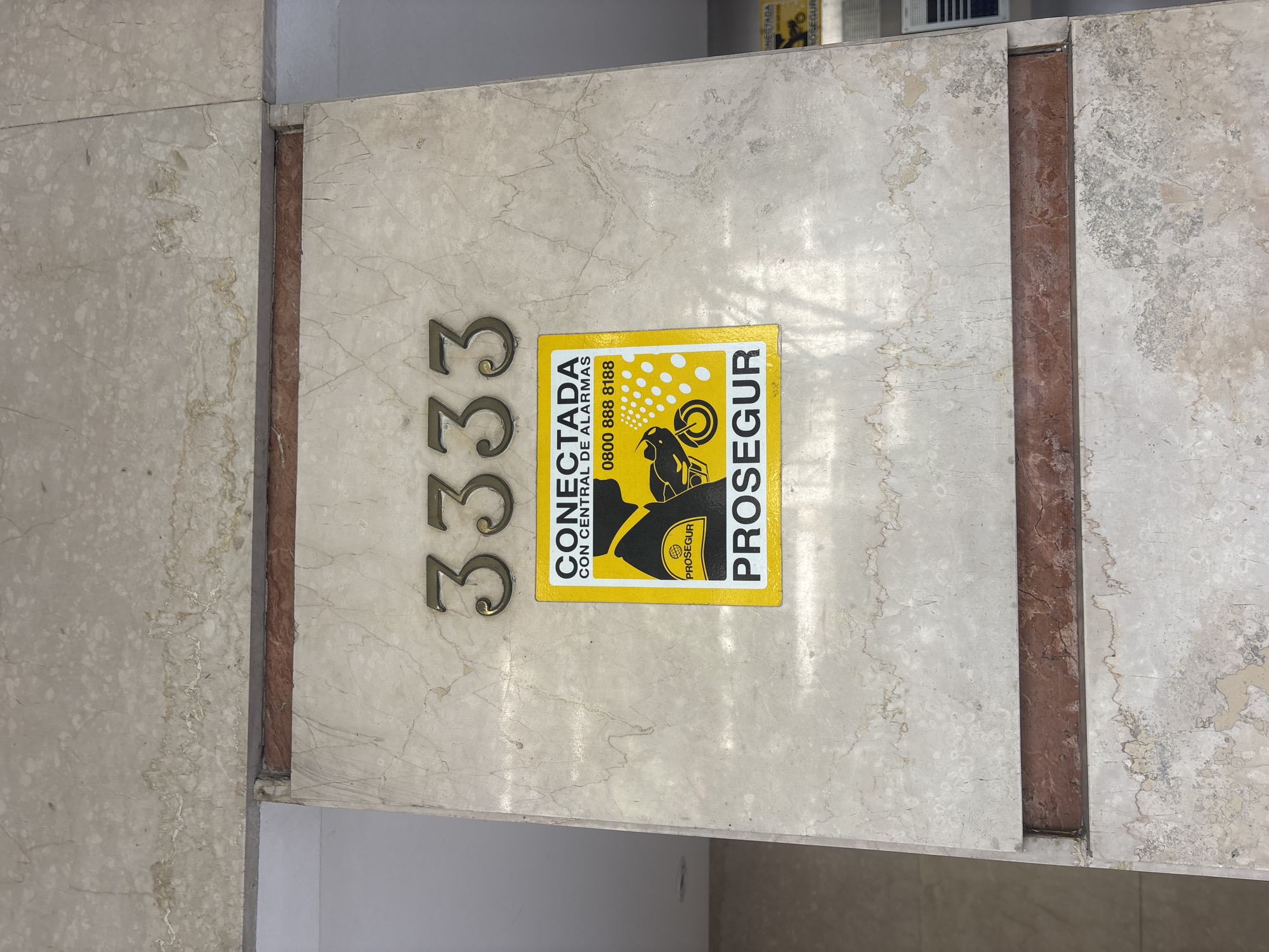 The image depicts a wall, likely the facade of a building, with the street number "3333" displayed vertically in bronze-colored numerals. A yellow and black security alarm sticker from the company "Prosegur" is affixed next to the numbers. The sticker reads "CONECTADA CON CENTRAL DE ALARMAS" and includes a phone number (0800 888 8188) and an image of a person in a protective suit riding a motorcycle in pursuit. The wall is constructed from marble-like stone slabs with reddish-brown columns on either side. The scene is in daylight.
Given the use of Spanish in the sticker text and the presence of Prosegur, this location is likely in Argentina. While the exact location in Buenos Aires cannot be determined from this image alone, it's evident that this is a building or property with Prosegur security alarm service.