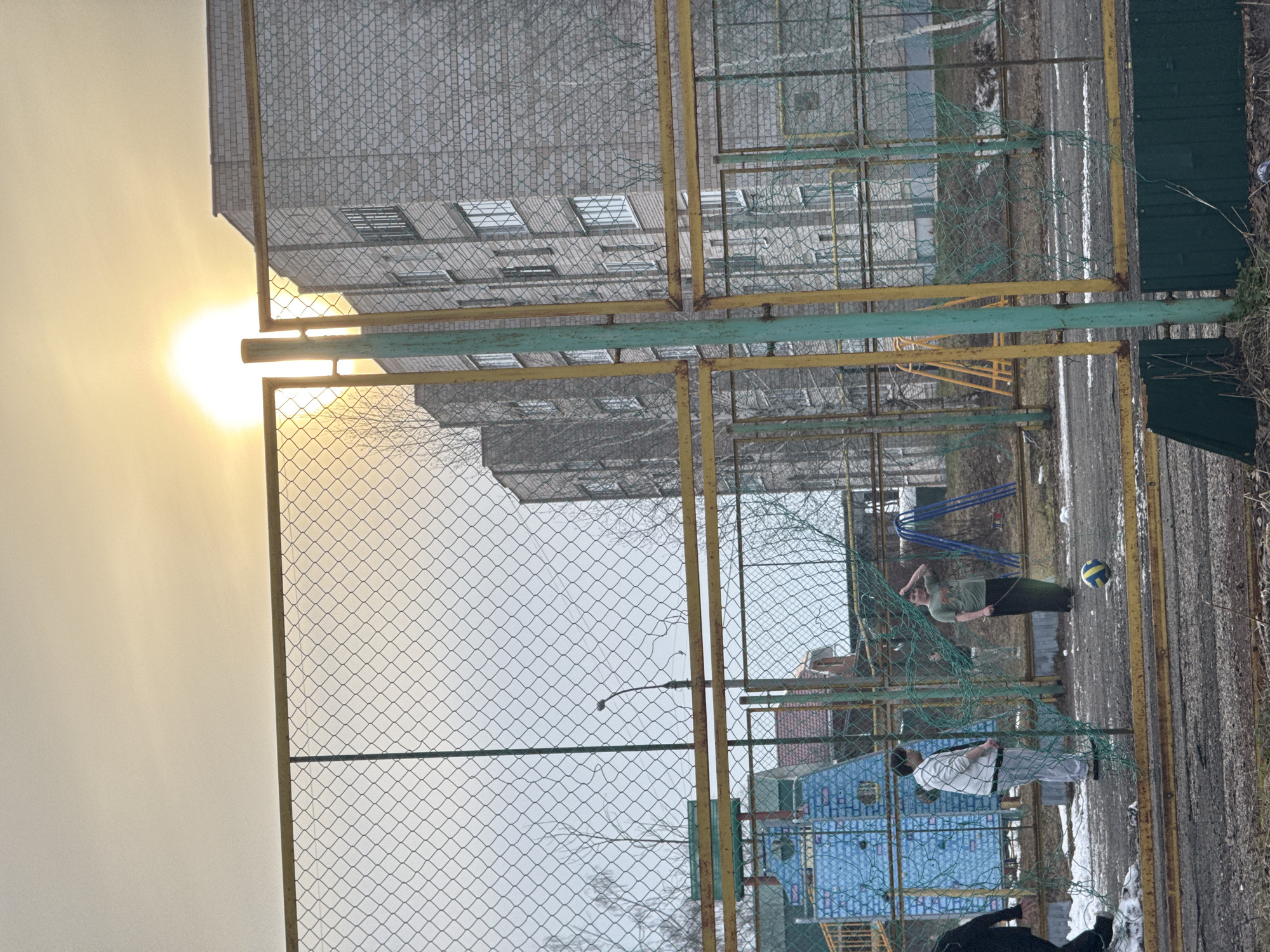 The photograph shows an outdoor volleyball/sports area located within the city of Votkinsk, Russia, as per image tags. The area is enclosed by a metal chain-link fence with a painted yellow frame. The image is taken from within the fenced area, facing the outside.
In the background is an apartment building with multiple windows. The sky appears overcast with the sun appearing partially obscured.
Inside the fence and to the right, two people are present. One is wearing a green shirt and black pants and is standing with their hand on their head as if shading their eyes or tired. A volleyball is positioned nearby. The other person wears a light-colored checkered shirt and light-colored pants, with some dark footwear, standing still. Another person is partially seen at the bottom of the image, wearing black clothing.
In the background behind the fence, other buildings and a playground can be seen. There is snow on the ground and what appears to be melting snow/slush on the surface of the sports area.