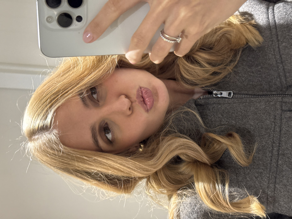 A woman with light blonde, wavy hair poses, holding a white smartphone up to take a selfie. She wears a gray zip-up jacket. Two rings adorn her hand holding the phone, and her fingernails are painted a light pink color. The picture is taken at a slight angle, with the woman's face tilted towards the camera and puckered lips. The background is a soft, blurred light color.