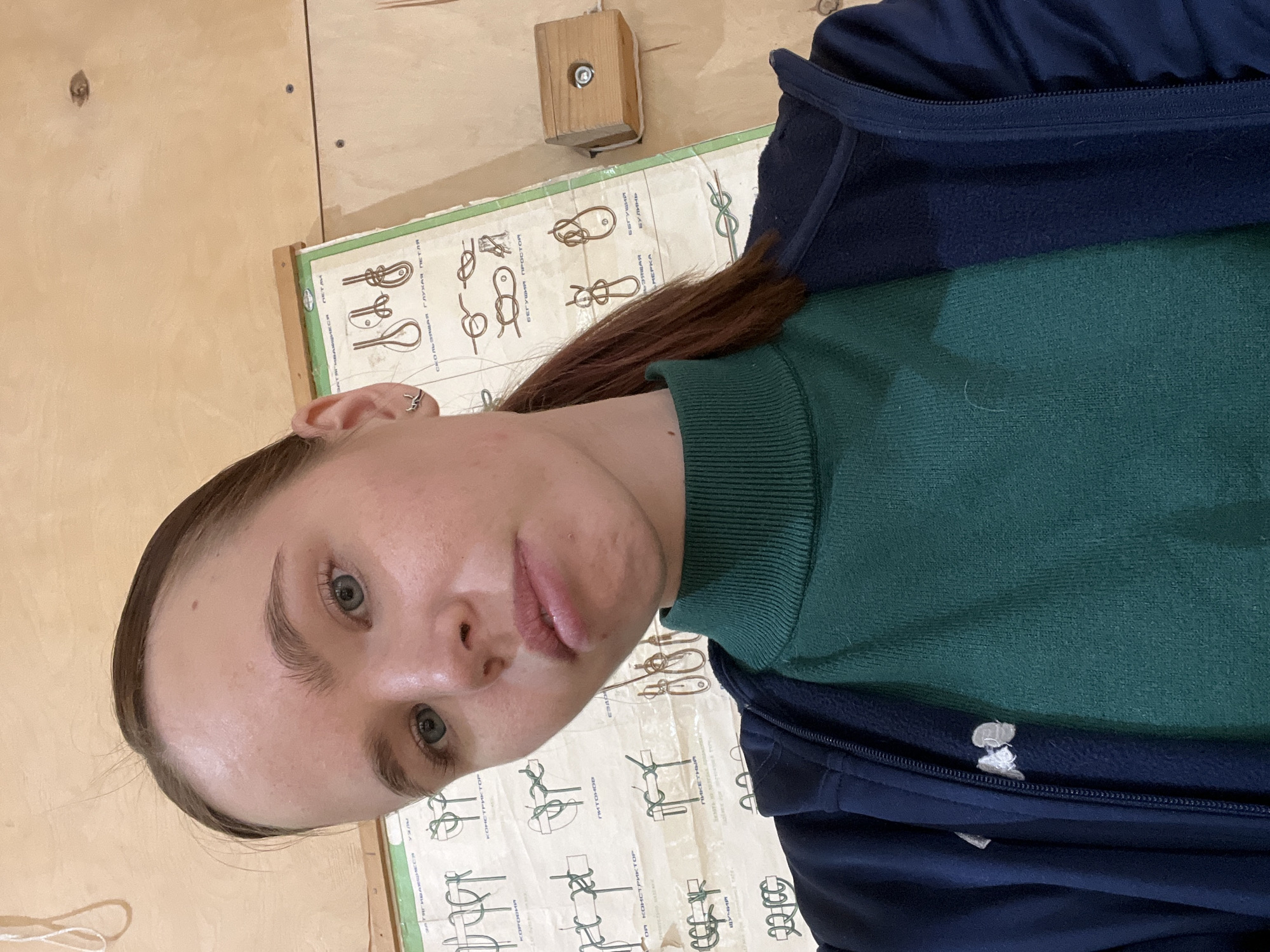 A person with light skin and brown hair pulled back in a ponytail is positioned in front of a poster depicting different types of knots. The person is wearing a green turtleneck sweater and a blue zip-up jacket. The poster is mounted on a wooden wall, with a small wooden block with a bolt attached to the wall above the poster. The text on the poster is in Russian.
There is no information available to determine the location as the city of Chaikovsky, Russia.