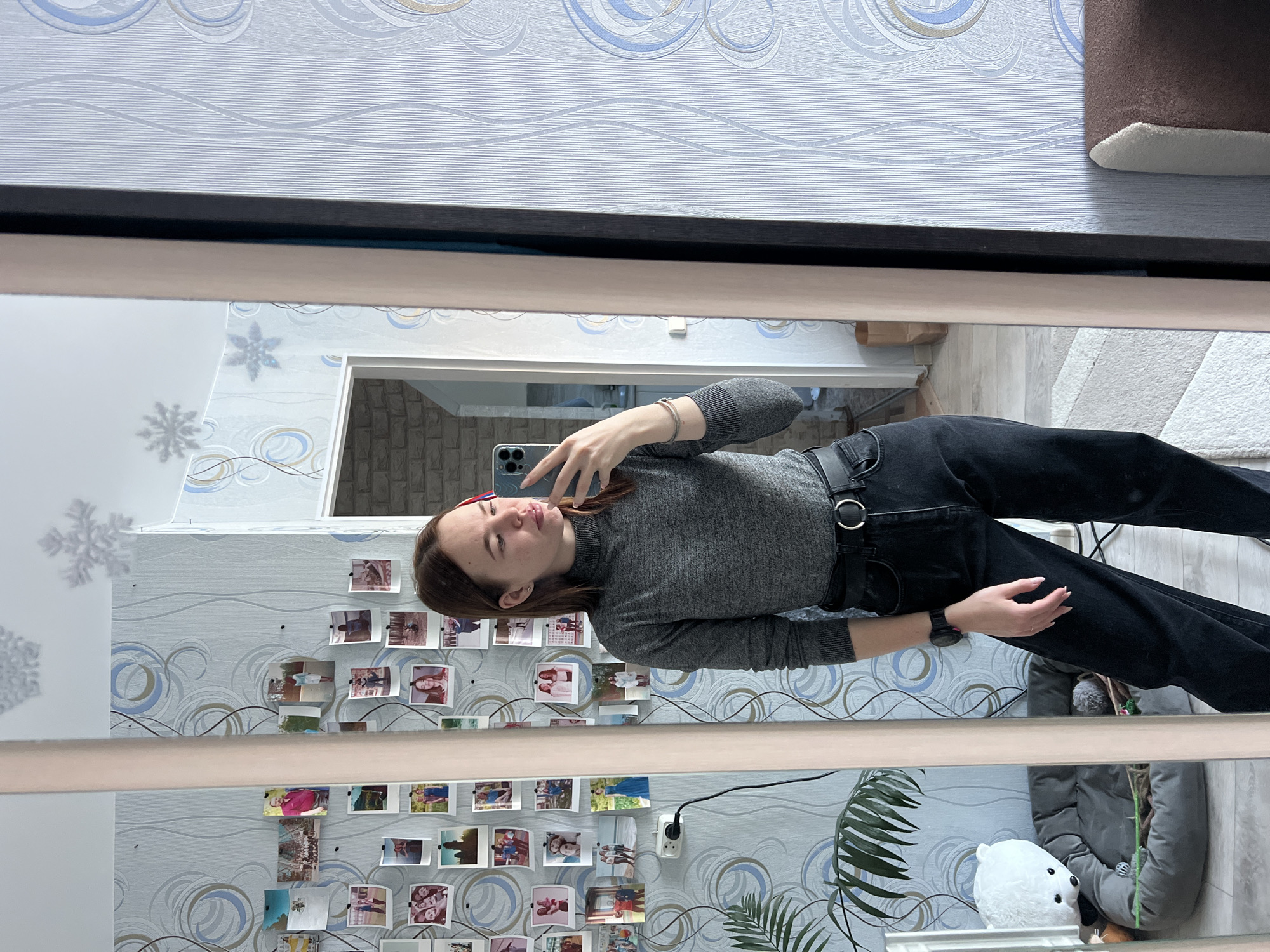A young woman is taking a selfie in a mirror. She is wearing a gray long-sleeved shirt, black pants, and a black belt. The mirror reflects a wall with wallpaper decorated with swirling patterns and many small photographs affixed to it. There is a plant and a white polar bear stuffed animal visible in the reflection as well. Based on other available information, this photo was taken in Chaikovsky, Russia.