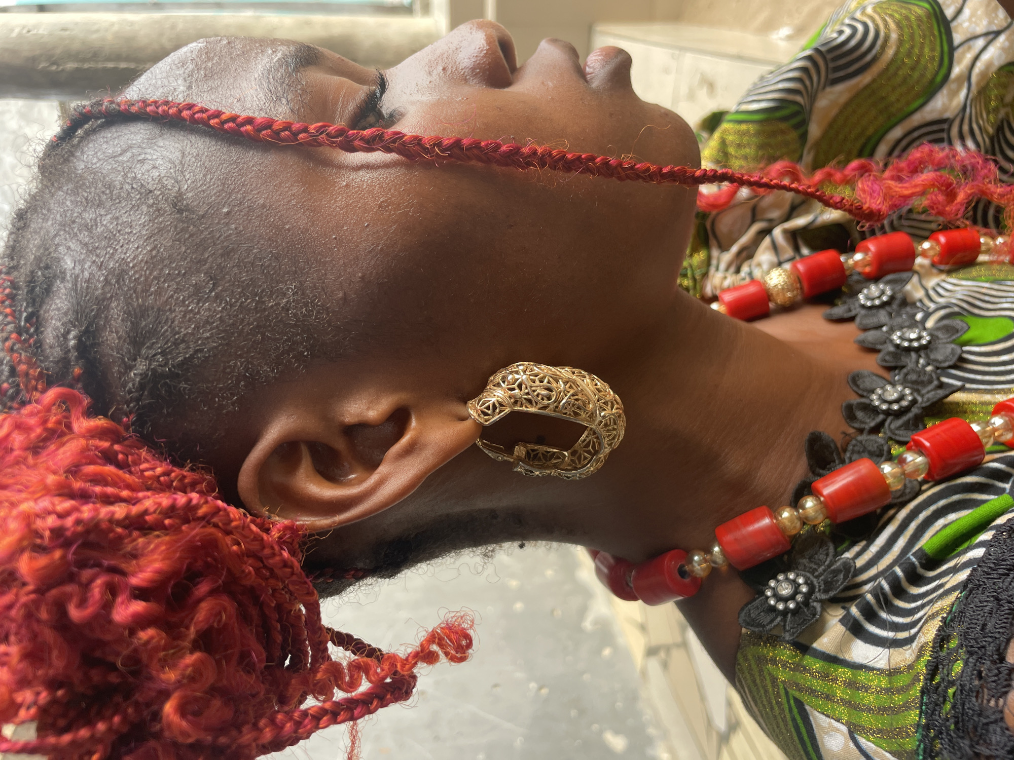A close-up, high-angle shot captures the right side of a person's head, neck, and shoulder, adorned with striking accessories. The individual's dark skin contrasts with their vibrant red, braided hair, which cascades around the ear and neck, some sections appearing tightly woven while others are loose and curly.
A prominent gold-toned hoop earring, intricately designed with an openwork filigree pattern, is worn on the right ear. Around the neck, a decorative necklace features large, cylindrical red beads interspersed with smaller, spherical gold or amber-colored beads. Black, floral-shaped fabric or beaded embellishments, some with small silver-toned centers, are also part of the necklace, resting on the shoulder and chest.
The person is wearing a garment with a lively pattern consisting of wavy lines in green, black, white, and hints of gold sparkle. The background is softly blurred and light-colored, suggesting an indoor or shaded outdoor setting. The bright, natural-looking light indicates it is likely daytime. The overall presentation suggests a deliberate adornment, possibly for a special occasion or cultural expression in Badagry, Nigeria. There is no visible text in the image.