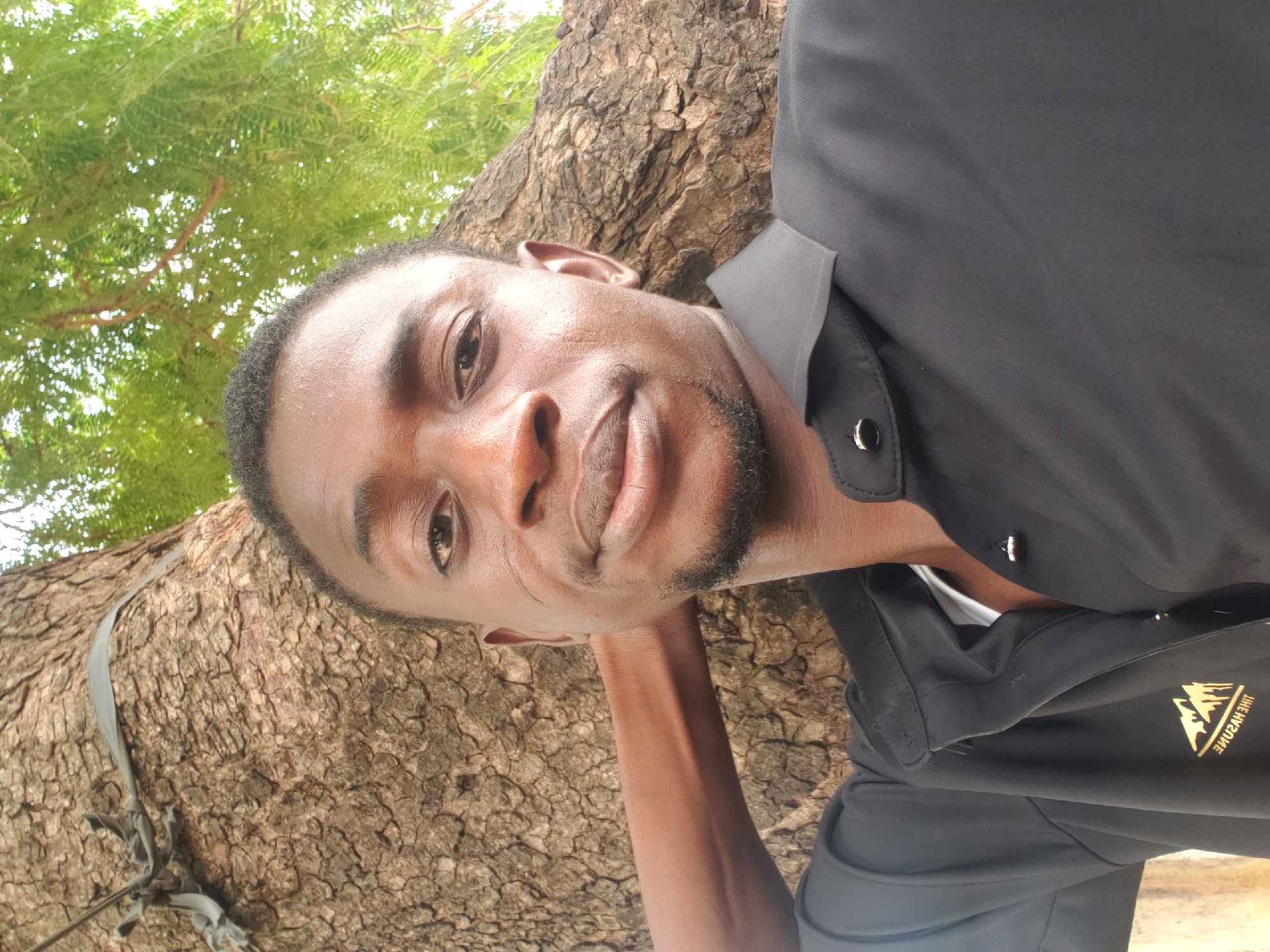 The image features a portrait of a young Black man in an outdoor setting. His head is tilted to the right, and he looks directly at the camera with a calm, slightly smiling expression. He has short dark hair and a neatly trimmed beard and mustache. He is wearing a black collared shirt, with a white undershirt subtly visible at the neckline, and his right arm is bent with his hand resting behind his head or neck.
The background is dominated by a large tree trunk with deeply textured, brown bark, extending diagonally from the lower left to the upper right of the frame behind the man. Lush green foliage, likely from the same tree, is visible in the upper portion of the image. The scene is illuminated by bright, natural daylight, suggesting a clear day, possibly late morning or early afternoon.
On the man's black shirt, a gold emblem is visible, depicting three mountain peaks. Below the mountains, text is partially visible, which, when deciphered from the angle, reads "THE HVNTER." The setting is consistent with an outdoor natural environment in Adankolo, Nigeria.