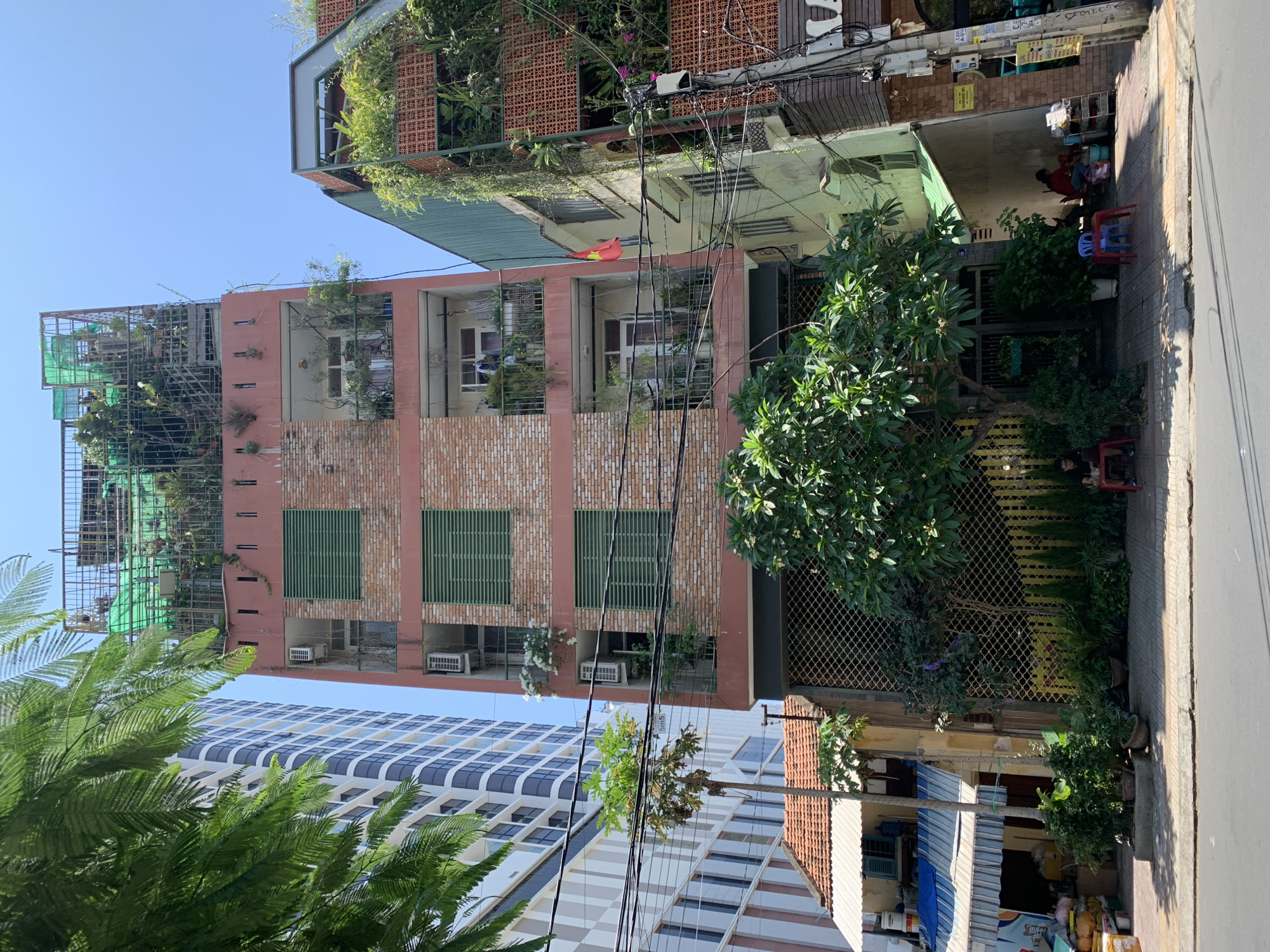 The image captures a vibrant street scene in an urban area under a clear blue sky, indicating bright daylight. In the mid-ground, a prominent multi-story building features a peach-colored facade with sections of light brickwork and green slatted windows. Its balconies and rooftop are densely covered with lush green plants and trees, some supported by metal frames, giving it the appearance of a vertical garden. Several air conditioning units are visible on its lower levels.
To the right of this building, an older, smaller structure with a dark green corrugated roof and orange-red patterned brickwork is seen. A large, leafy tree with small white flowers stands in front, partially obscuring a metal gate. On the tiled sidewalk in front of this building, a person is seated on a small red plastic stool, facing away, next to what appears to be a small street stall with various items. Another person is partially visible near the base of the large tree. Further down the street, another building with a red-tiled roof and a blue-and-white striped awning is visible.
In the background, a modern high-rise building with a distinctive blue and white grid-patterned facade stands tall. In the immediate foreground, large green fronds from a palm-like tree partially obscure the view. Overhead, numerous black electrical wires crisscross the sky. A notable detail is a red and yellow bird-like sculpture or decoration perched on one of the wires.
Visible text includes "Bún Đậu" on a sign and "TRA" or "Trà" (Vietnamese for tea) on a blue and white striped awning. Another small sign displays "5m5". The scene depicts typical daily life, blending traditional and modern architecture with abundant greenery.