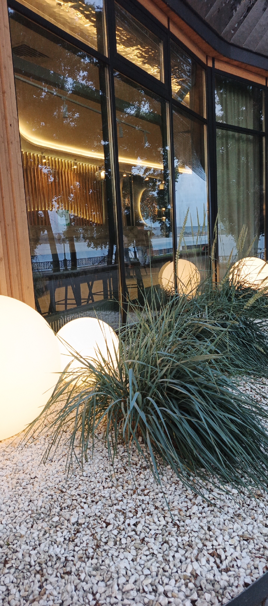 This is a low-angle, eye-level, vertical outdoor shot of a building with large windows, likely in the city of Votkinsk, Russia. The building has a modern design with wooden accents on the left side and dark-framed windows. Inside the building, visible through the glass, are vertical wooden slats illuminated by warm, yellow light, and a curved, art-deco-like light fixture. Reflections in the windows show trees and a body of water, possibly the Votkinsk Reservoir, with a fence and streetlights along the bank.
In the foreground, a patch of light-colored gravel is covered by the base of ornamental grass. Two large, illuminated spherical outdoor lights, glowing with a soft white light, are partially visible behind the grass on the left. The overall scene suggests an evening or dusk setting, highlighting the interplay of light and reflection. There are no people clearly visible within the frame.
