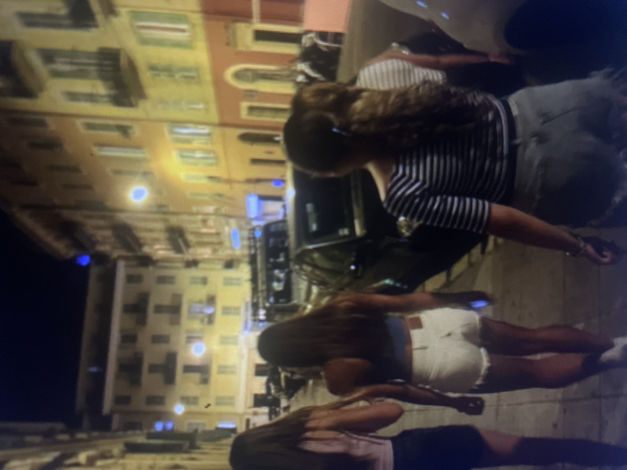 Four individuals, appearing to be young women, are walking down a cobblestone street at night. The architecture of the buildings lining the street suggests a European city, with multiple stories and balconies. The lighting is dim, with streetlights illuminating the scene. The individuals are dressed in casual summer attire. The woman in the foreground is wearing a horizontally striped shirt and denim shorts. The woman to her left is wearing white denim shorts and a dark tank top. Another woman to the far left is wearing dark shorts and a light-colored top. A fourth individual, partially obscured, is visible behind the woman in the striped shirt. Several parked cars are visible on the street, and one car appears to be moving in the background. The overall impression is of a group enjoying an evening out in an urban setting. There is no specific information in the image to indicate the location is Kołobrzeg, Poland.