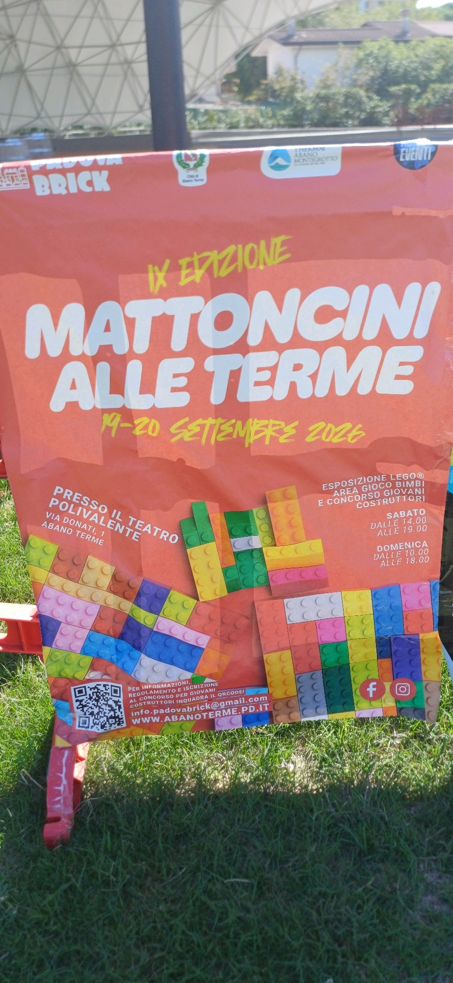 This is a photograph of a large, orange banner displayed outdoors. The banner advertises an event called "MATTONCINI ALLE TERME," which translates to "Bricks at the Spa." The event is described as the "IX EDIZIONE" (9th edition) and is scheduled for "19-20 SETTEMBRE 2026" (September 19-20, 2026).
The banner features graphics made to look like LEGO bricks in various colors, covering a significant portion of its surface. Text on the banner indicates the event is held at "PRESSO IL TEATRO POLIVALENTE" (At the Multipurpose Theater) located at "VIA DONATI, 1 ABANO TERME."
Additional details on the banner include "ESPOSIZIONE LEGO® AREA GIOCO BIMBI E CONCORSO GIOVANI COSTRUTTORI" (LEGO® Exhibition, Children's Play Area, and Young Builders Competition). It also lists the event hours: "SABATO DALLE 14.00 ALLE 19.00" (Saturday from 2:00 PM to 7:00 PM) and "DOMENICA DALLE 10.00 ALLE 18.00" (Sunday from 10:00 AM to 6:00 PM).
Contact information is provided, including a QR code, an email address "info.padovabrick@gmail.com," and a website "WWW.ABANOTERME.PD.IT." Social media icons for Facebook and Instagram are also visible.
The banner is supported by a red structure and is set against a background of green grass, suggesting it is located in an outdoor public space. In the upper part of the image, a white tent-like structure and some trees and buildings are visible, indicating a daytime outdoor setting. The lighting suggests the sun is present, casting shadows. There are no people clearly visible in the frame.