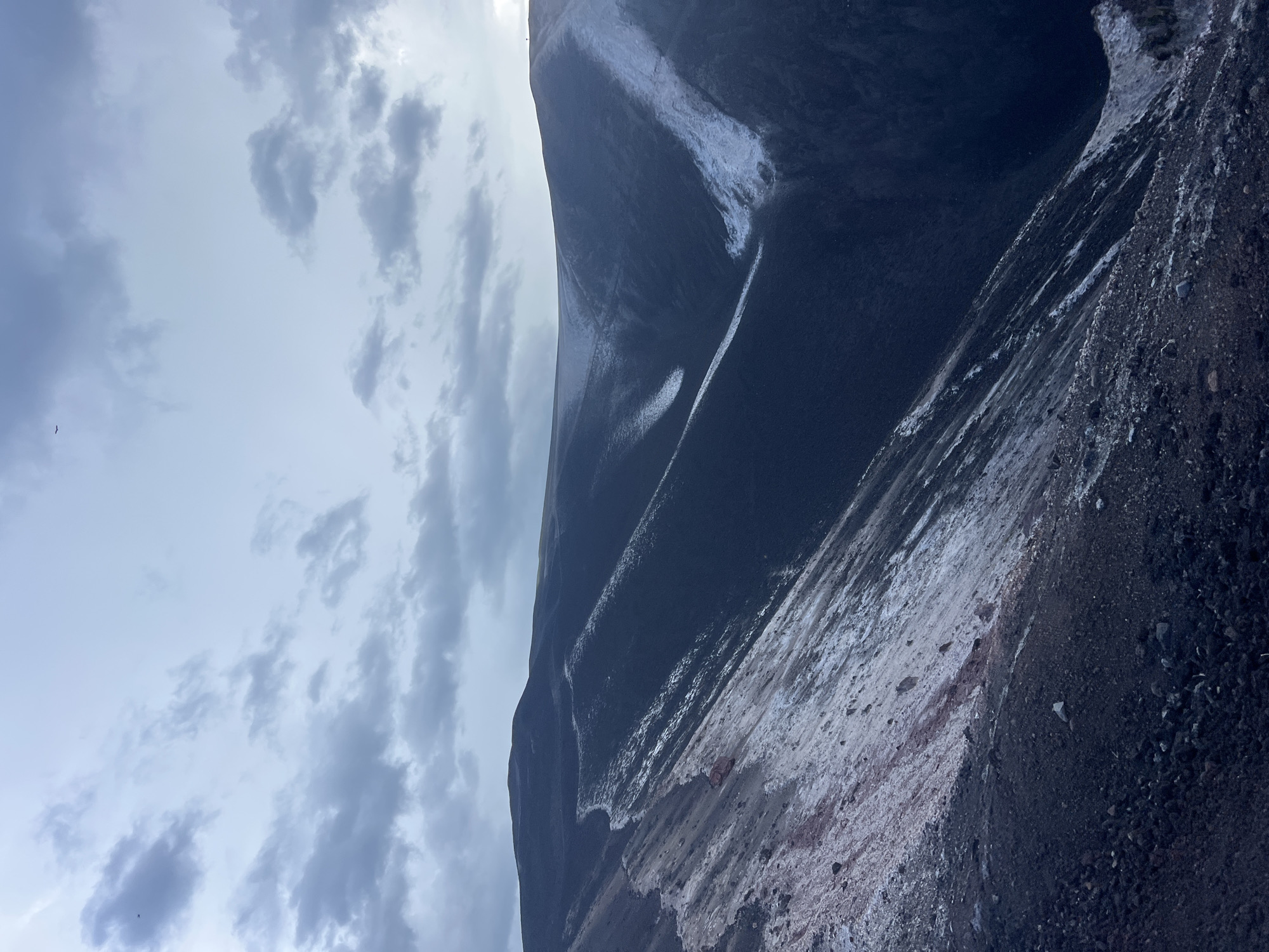 The image shows the rim of a volcanic crater. The ground is covered in dark volcanic rock and ash. Patches of white, possibly snow or mineral deposits, are visible on the slopes. The sky is cloudy and overcast. There are no people or visible man-made structures in the image. The scene is a natural landscape.