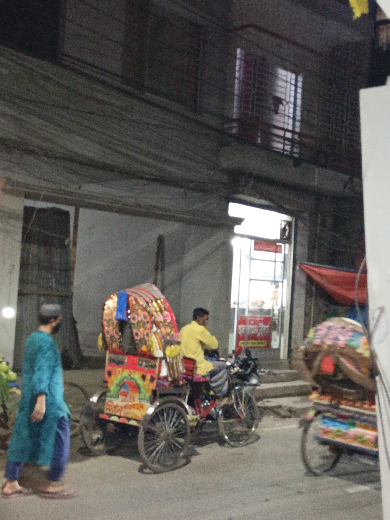 This low-light, blurry image depicts a nighttime street scene in an urban area of Dhaka, Bangladesh, characterized by overhead wires and dim artificial lighting.
In the foreground, a person wearing a teal tunic, blue pants, and a skullcap walks towards the right. Alongside them is a vibrant cycle-rickshaw, a prevalent form of transport in Bangladesh. The rickshaw's canopy and rear body are elaborately decorated with colorful, folk-art inspired paintings, including a house and landscape. Unreadable Bengali text is also visible as part of the rickshaw's adornment. A man in a yellow shirt is seated on this rickshaw, facing away from the camera. Another similarly decorated cycle-rickshaw is partially visible on the far right, heading out of frame.
The background features a multi-story building. Its upper floor has windows secured with metal grilles, one of which has a distinct red metal railing. Below, a brightly lit storefront with a glass door reveals red and white signs inside, bearing unreadable Bengali text. The street itself appears to be a paved road, leading up to steps at the storefront entrance. The scene captures typical urban activity and architecture of the region during evening hours.