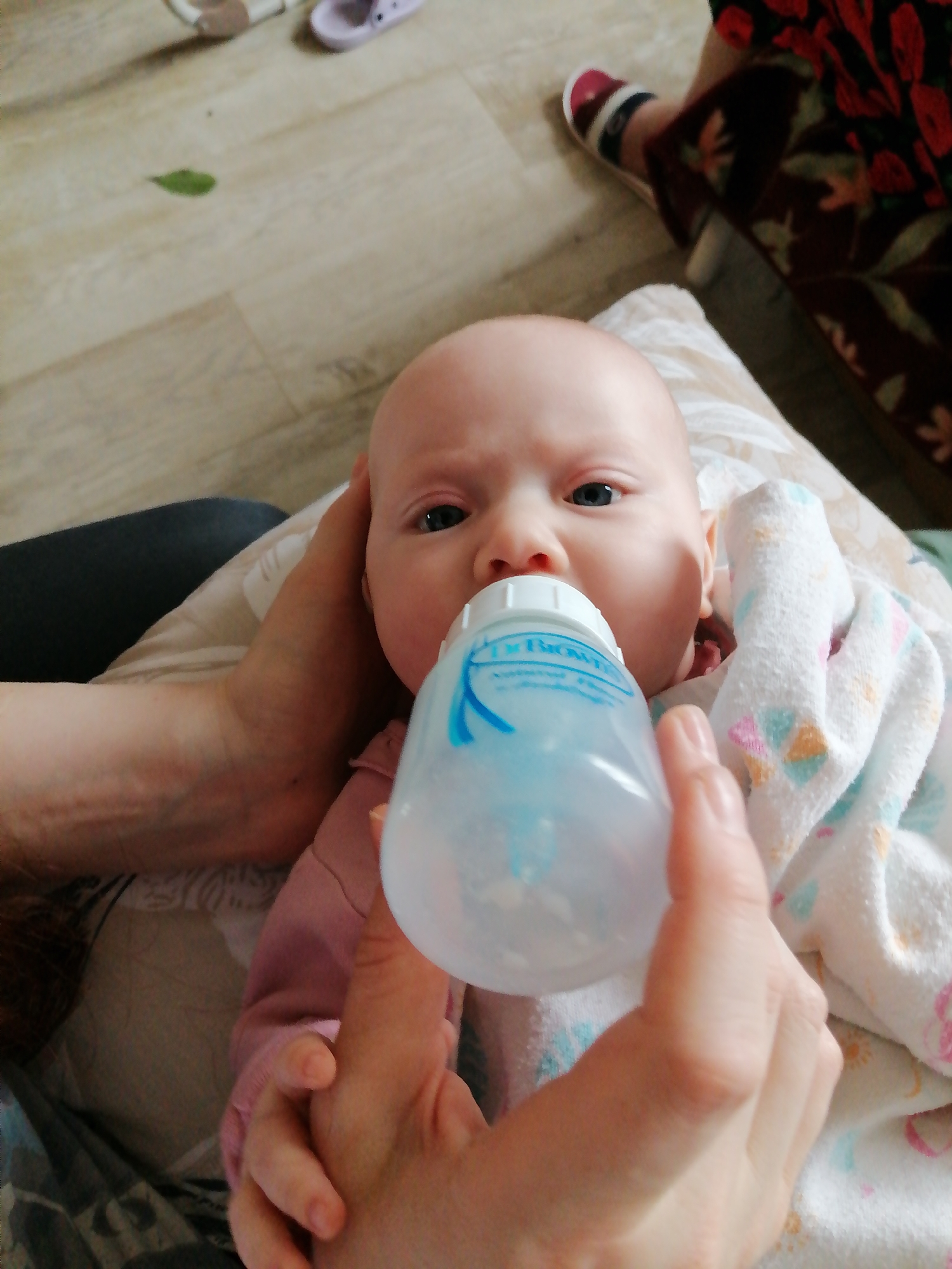 A baby is being bottle-fed indoors. The baby is lying on a patterned surface, possibly a cushion or blanket. An adult hand supports the baby's head, while another holds a Dr. Brown's baby bottle to the baby's mouth. The baby is wearing a pink top and is partly covered by a white blanket with pastel-colored patterns. In the background, there's a glimpse of flooring, footwear, and a person wearing a patterned dress. The location appears to be inside a residential home in the city of Unknown, Unknown.
