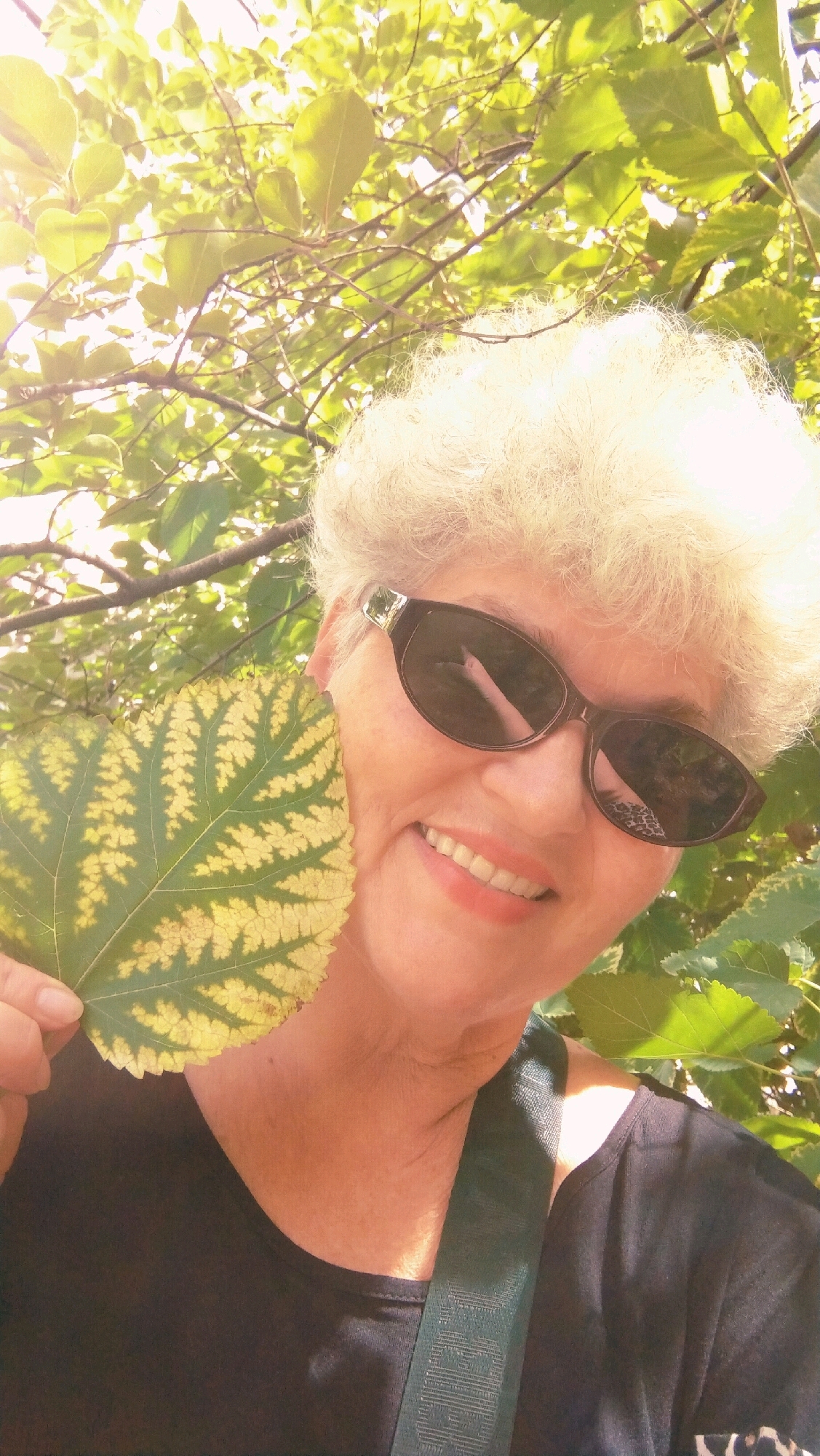 A woman with short, blonde, permed hair and sunglasses is holding a green and yellow leaf in front of her face. She is smiling broadly, and her teeth are visible. She is wearing a black shirt with a green strap across her chest. The background is filled with green leaves and sunlight filtering through the trees, suggesting an outdoor setting, possibly a park or garden in Horishni Plavni, Ukraine.