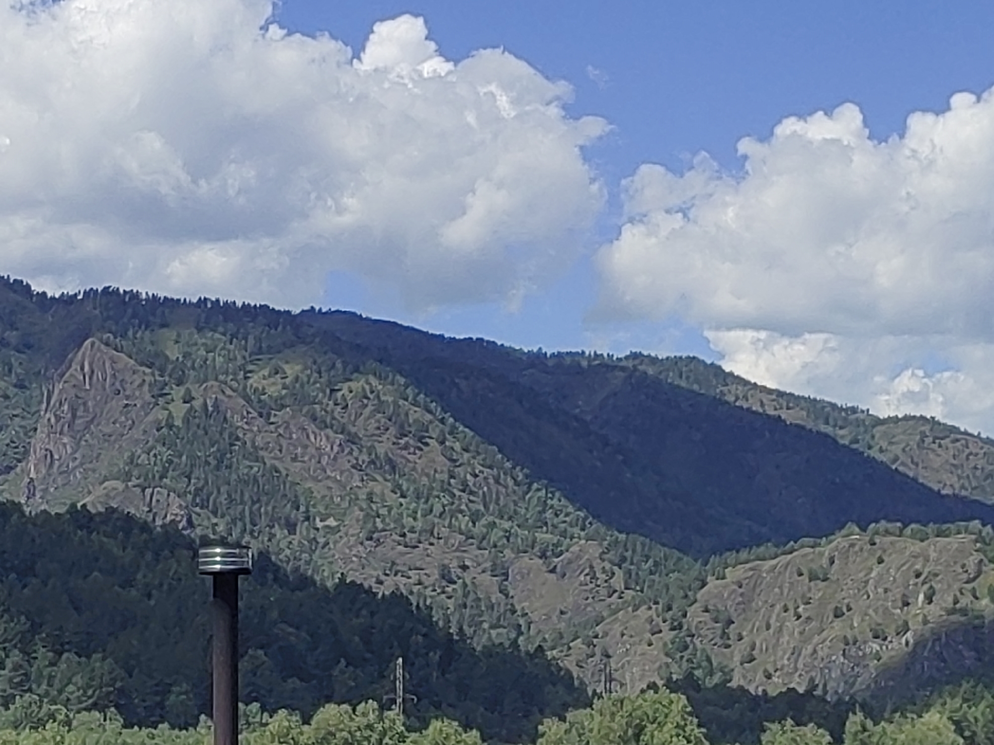 The image shows a mountainous landscape with a clear blue sky filled with fluffy white clouds. The mountains are covered with lush green trees and vegetation, with rocky outcrops visible on some slopes. In the foreground, a dark brown pole with a silver, cylindrical object on top stands prominently. This object appears to be some sort of sensor or light fixture. In the background, several layers of mountains recede into the distance, their slopes covered in a dense forest. The overall impression is one of a serene and natural environment, possibly a hiking or scenic viewpoint in the mountainous region of Chemal, Russia.