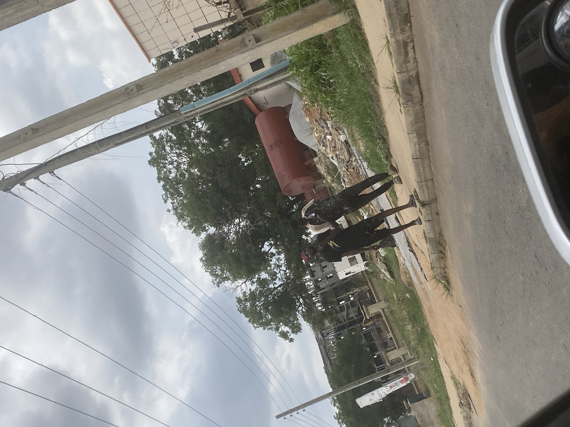 The scene is a street in Lugbe, Nigeria, viewed from within a vehicle. Two individuals are walking along the roadside. A large, reddish-brown cylindrical tank is positioned near a building under construction. The environment includes greenery, buildings in varying states of completion, overhead power lines, and a cloudy sky. A portion of the vehicle's side mirror is visible.