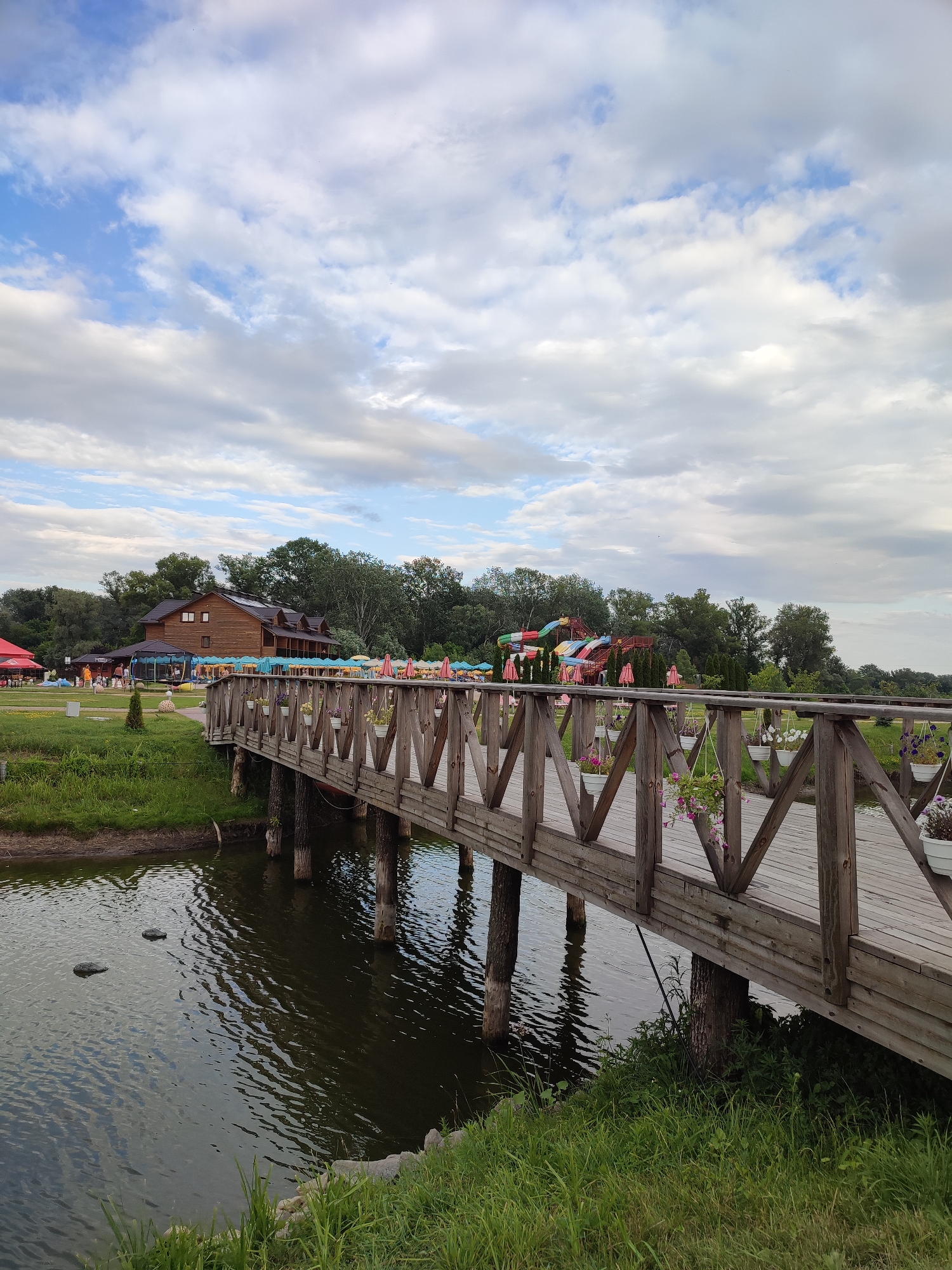 This is a medium shot photograph of a wooden footbridge crossing a dark body of water. The bridge is constructed of rough-hewn wooden planks and supported by thick, vertical wooden posts. The railings of the bridge are made of diagonal wooden slats, forming a geometric pattern. Hanging flower pots with pink and white blossoms are attached to the outer side of the bridge railing.
Beyond the bridge, a large, two-story wooden building with a dark roof and multiple windows is visible. Several umbrellas, some red and some pink, are scattered across the grassy area in front of the building. To the left of the building, a red tent is pitched. In the background, a cluster of colorful water slides stands out against a backdrop of lush green trees. The sky above is cloudy, with patches of blue peeking through.
The scene appears to be a recreational area or a park. The overall ambiance is one of a peaceful, outdoor setting on a cloudy day. The photograph was taken in the city of Shcherbani, Ukraine.