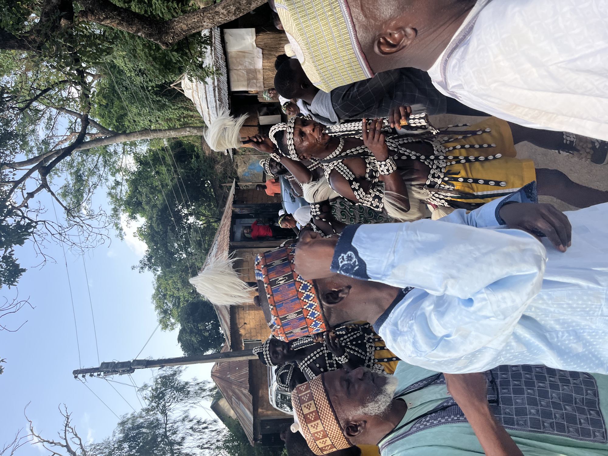 The image shows a group of men in what appears to be an outdoor gathering in Jos, Nigeria. The primary subjects are several men dressed in traditional attire. One man in the foreground wears a light blue garment and a patterned cap, with a white beard. Behind him, another man is adorned with a white headdress, a necklace of shells and beads, and bracelets. He is holding a white whisk or fan. Other men are visible in the background, also in traditional clothing, some with similar headdresses and ornamentation.
The scene suggests a cultural event or ceremony, possibly a festival or a traditional gathering. The setting is somewhat rural or semi-urban, with buildings with corrugated iron roofs visible, along with trees and vegetation. A utility pole with wires is also present, indicating the presence of infrastructure. The lighting suggests it is daytime, likely under a clear sky, as indicated by the bright light and shadows.
The expressions on the faces of the men are difficult to discern clearly due to the angle and focus, but there seems to be a sense of participation and engagement in the event. The ornate attire and accessories point to a culturally significant occasion. There is no visible text in the image.