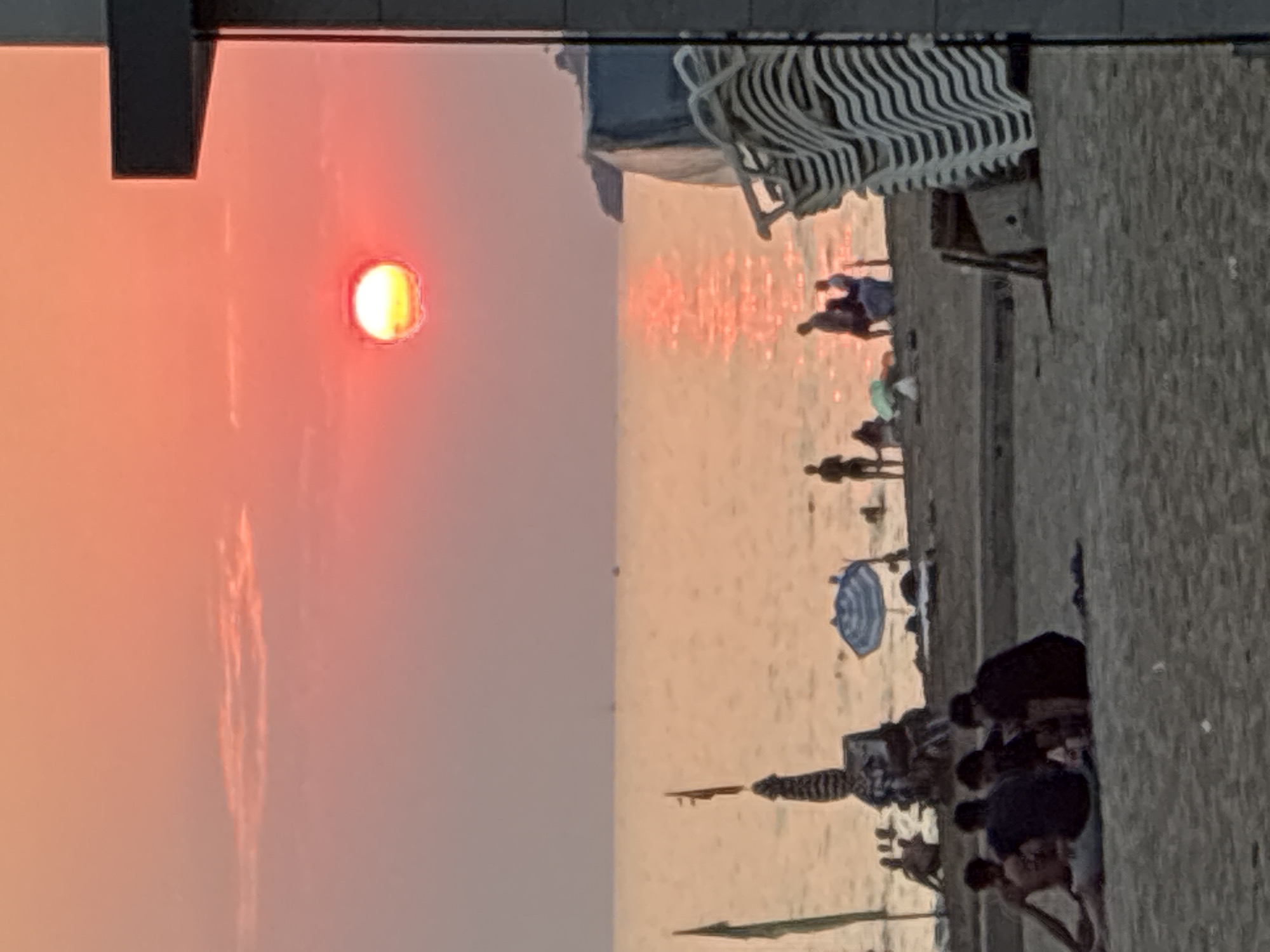 A vibrant sunset paints the sky in hues of pink and orange over the sea in Alanya, Turkey. The sun, a bright red orb, is positioned in the upper right quadrant of the frame, its light reflecting on the water. On the sandy beach in the foreground, a group of people are seated, some appearing to be engaged in conversation. Further down the beach, other figures are visible, some standing and others sitting, with beach umbrellas and chairs scattered around. The scene depicts a typical evening at a seaside resort.