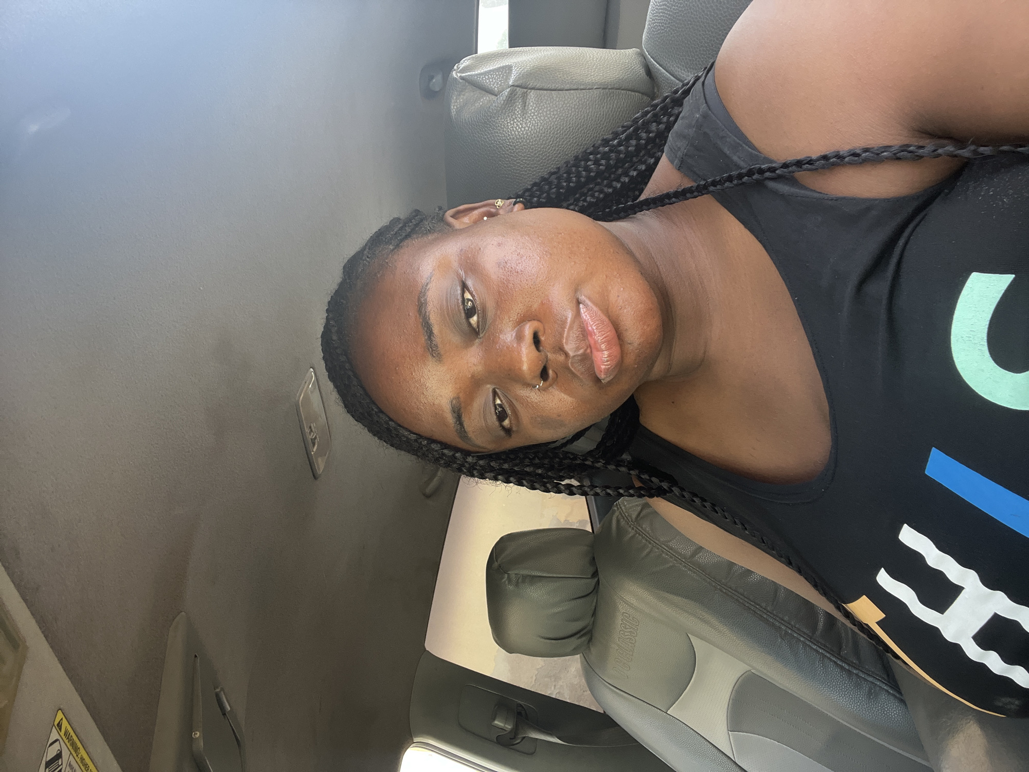 A young Black woman is depicted in a close-up, tilted shot, positioned inside what appears to be a car, likely in the passenger seat. She has long, dark box braids, a septum piercing, and a small gold hoop earring in her left ear. Her skin is clear, and she has full lips and dark eyes, looking directly at the camera with a neutral expression.
She is wearing a black tank top or t-shirt featuring a colorful graphic design on the front, which includes a light green 'C' shape, a blue rectangular element, and white wavy lines.
The car's interior is visible, with gray and black leather or vinyl seats, and a light gray fabric headliner. A "CLASSIC" label is partially embossed on the car seat. Through the car window, a bright, possibly dry or dusty, outdoor environment is discernible, suggesting a sunny day. A sun visor is visible in the upper part of the frame, with a partially obscured "WARNING: HIGHER RISK" label. The bright natural lighting indicates it is daytime. The image captures a casual moment, possibly a selfie.