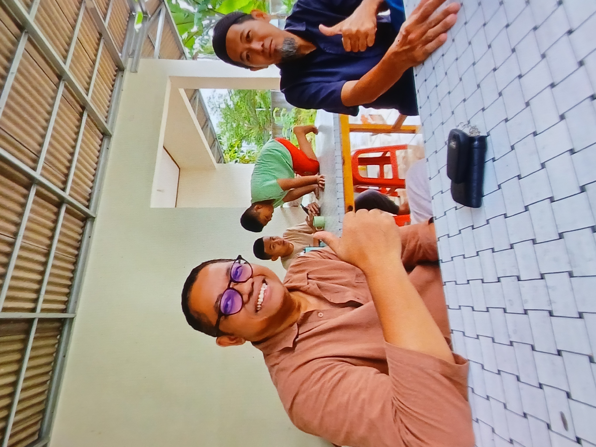 In Setia Bakti, Indonesia, a group of four individuals, three adults and one child, are gathered outdoors, seated and kneeling at a table. The scene is captured from a slightly low angle, looking up towards the subjects.

In the foreground, two men are positioned. The man on the left, wearing glasses and a brown collared shirt, smiles broadly and gives a thumbs-up gesture. He is seated at a table with a white and grey patterned surface. To his right, another man, wearing a dark blue shirt and a black cap, also gives a thumbs-up. He has a grey beard and is looking towards the camera.

In the background, a young boy in a green t-shirt and red shorts is kneeling on the table, looking down at a mobile phone. Another individual, possibly a boy or young man wearing a light brown shirt, is seated in the mid-ground, also appearing to be looking at a phone. Red stools are visible beneath the table.

The environment suggests an outdoor dining or gathering area with a corrugated metal roof supported by metal beams overhead. A wall with a pale green hue forms the background. Beyond the immediate area, lush green foliage and trees are visible, indicating a tropical or subtropical setting. A black wallet and keys are placed on the table in the foreground, to the right of the man in the brown shirt.