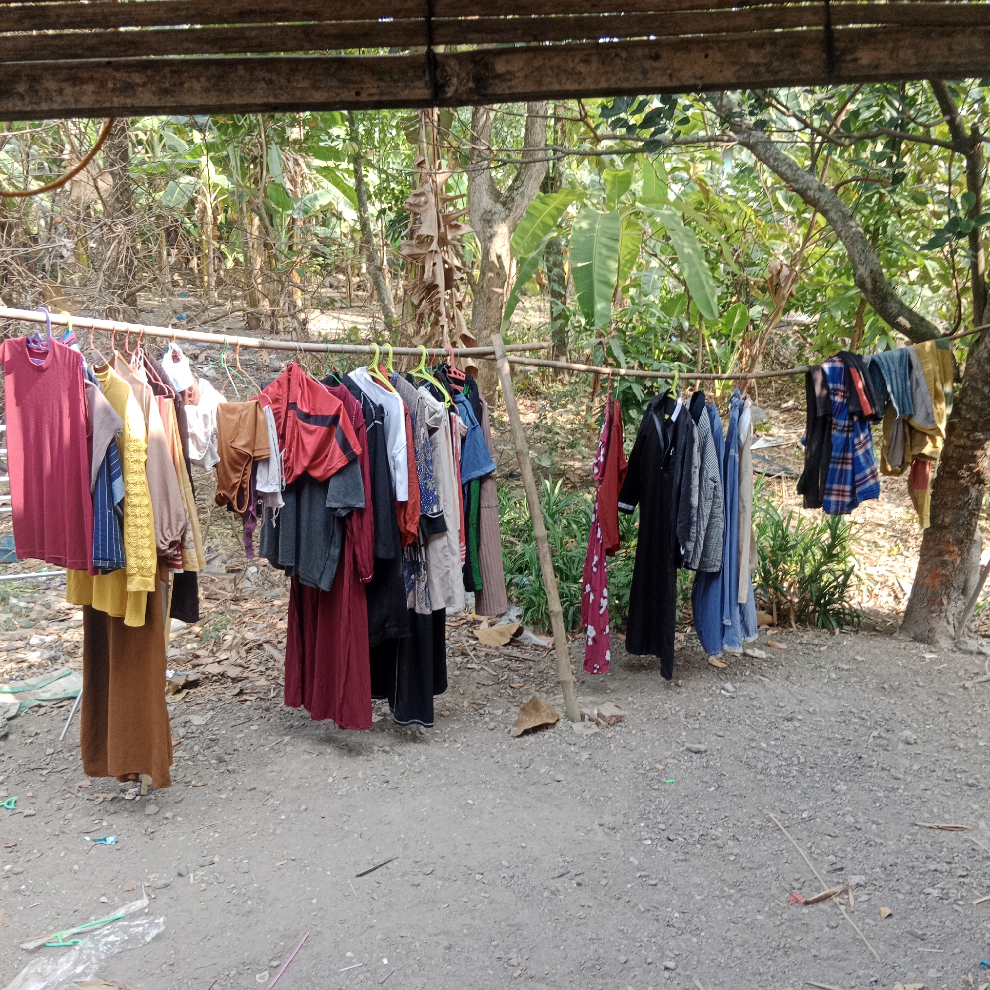 In Tegowanu Kulon, Indonesia, laundry is hung on a makeshift clothesline outdoors. The clothesline is supported by bamboo poles and a wooden structure overhead. Various items of clothing are displayed, including t-shirts, dresses, and pants, in a variety of colors and patterns. The ground is covered with gravel and dry leaves, and the background features lush green foliage from trees and plants. The scene suggests a rural or semi-rural setting.