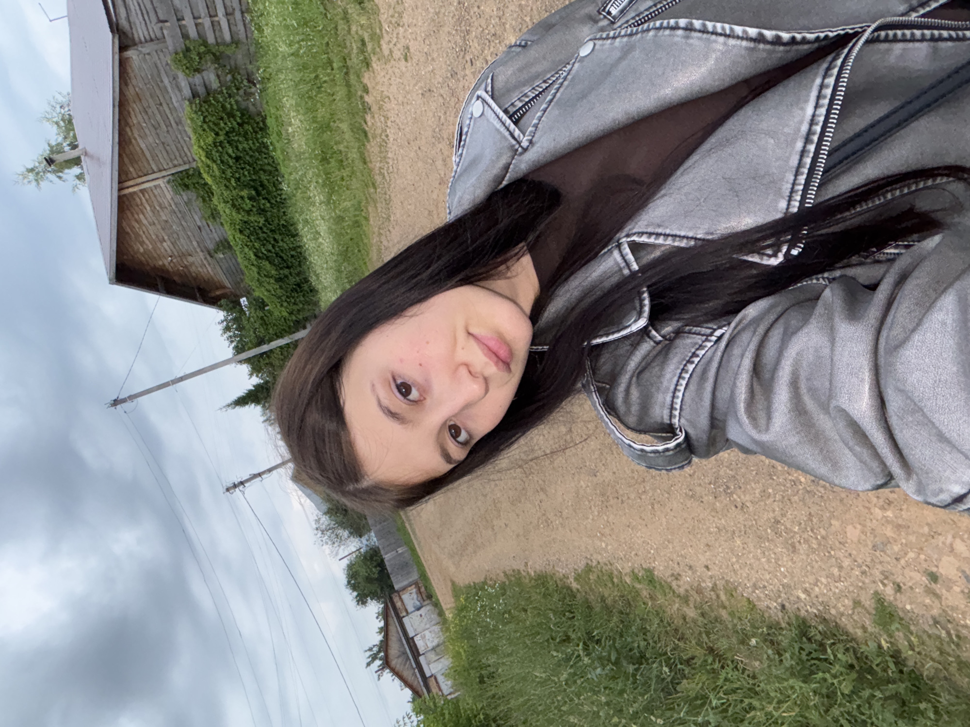 A young woman with long, dark hair is taking a selfie outdoors on a dirt road in Fokino, Russia. She is wearing a gray jacket over a brown shirt. To her right is a wooden building with a metal roof, partially covered in green foliage. To her left, the dirt road continues, bordered by tall green grass and wildflowers. In the background, there are more buildings and utility poles. The sky is overcast.