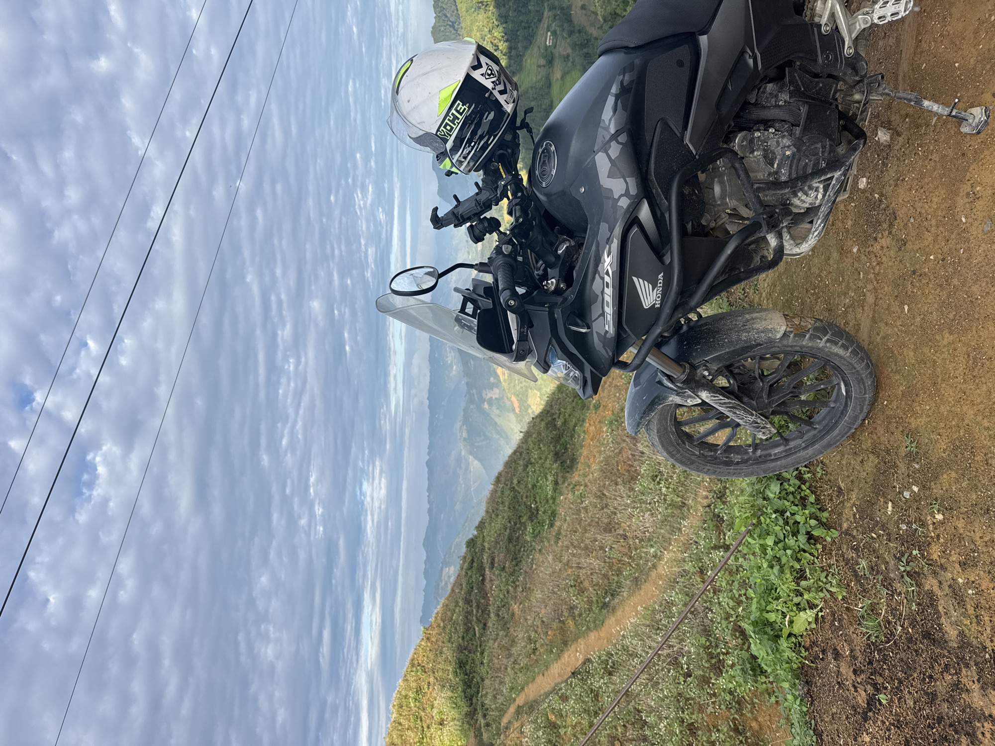 The image shows a black and grey Honda 500X motorcycle parked on a dirt path. A white and green helmet sits on the motorcycle's handlebars. The background reveals a mountainous landscape with cloudy skies. The photo was taken in Lũng Cú Commune, Vietnam.
