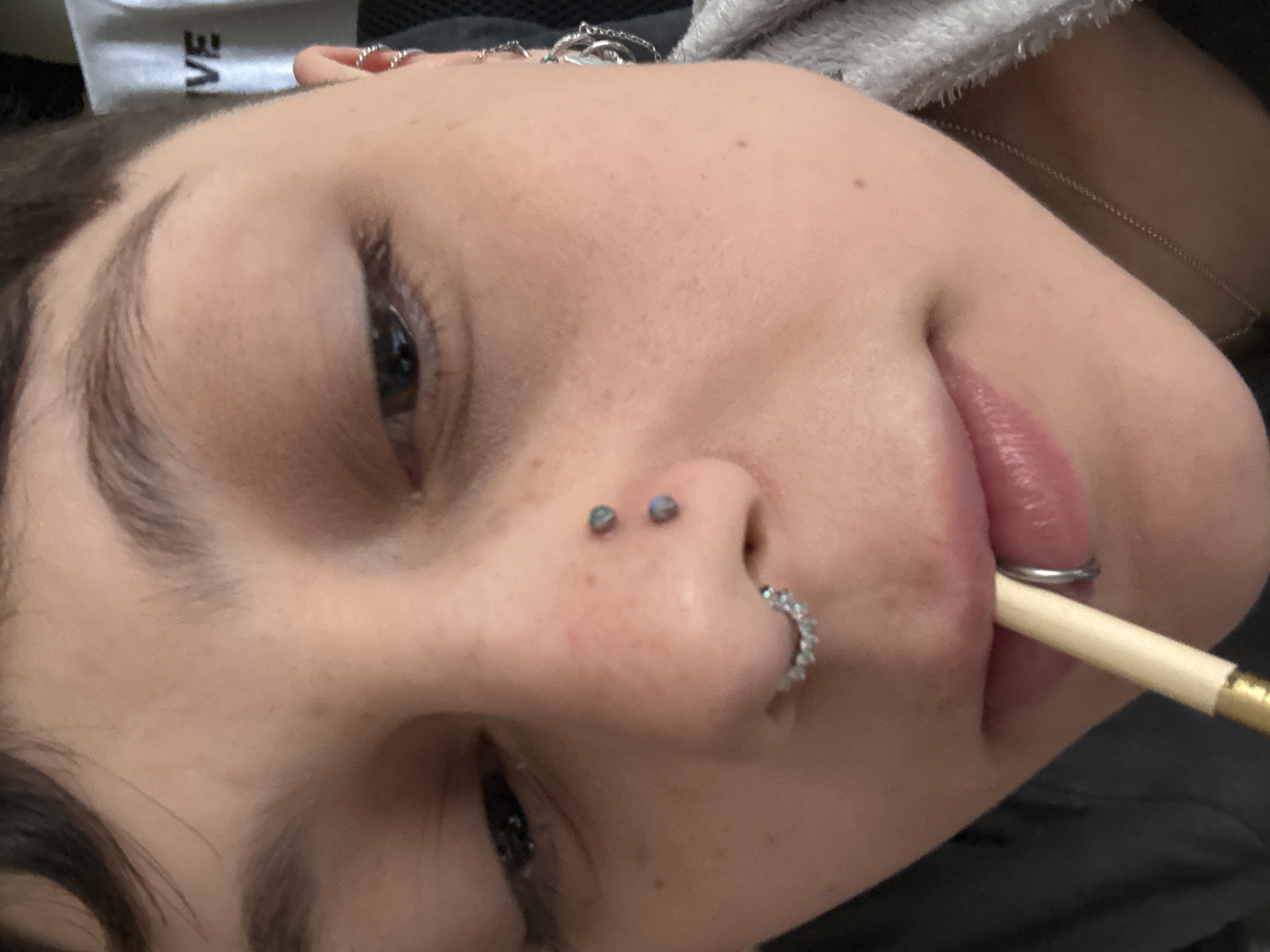 A person is lying down with their head tilted. The person has fair skin, dark eyebrows, and dark eyes. They have a double nose piercing on the left side of their nose and a ring piercing in the nostril on the right side. A cigarette is held between their lips. A silver chain is visible on their neck. The setting is indoors. There is no evidence or indication that this photo was taken in Moscow, Russia.