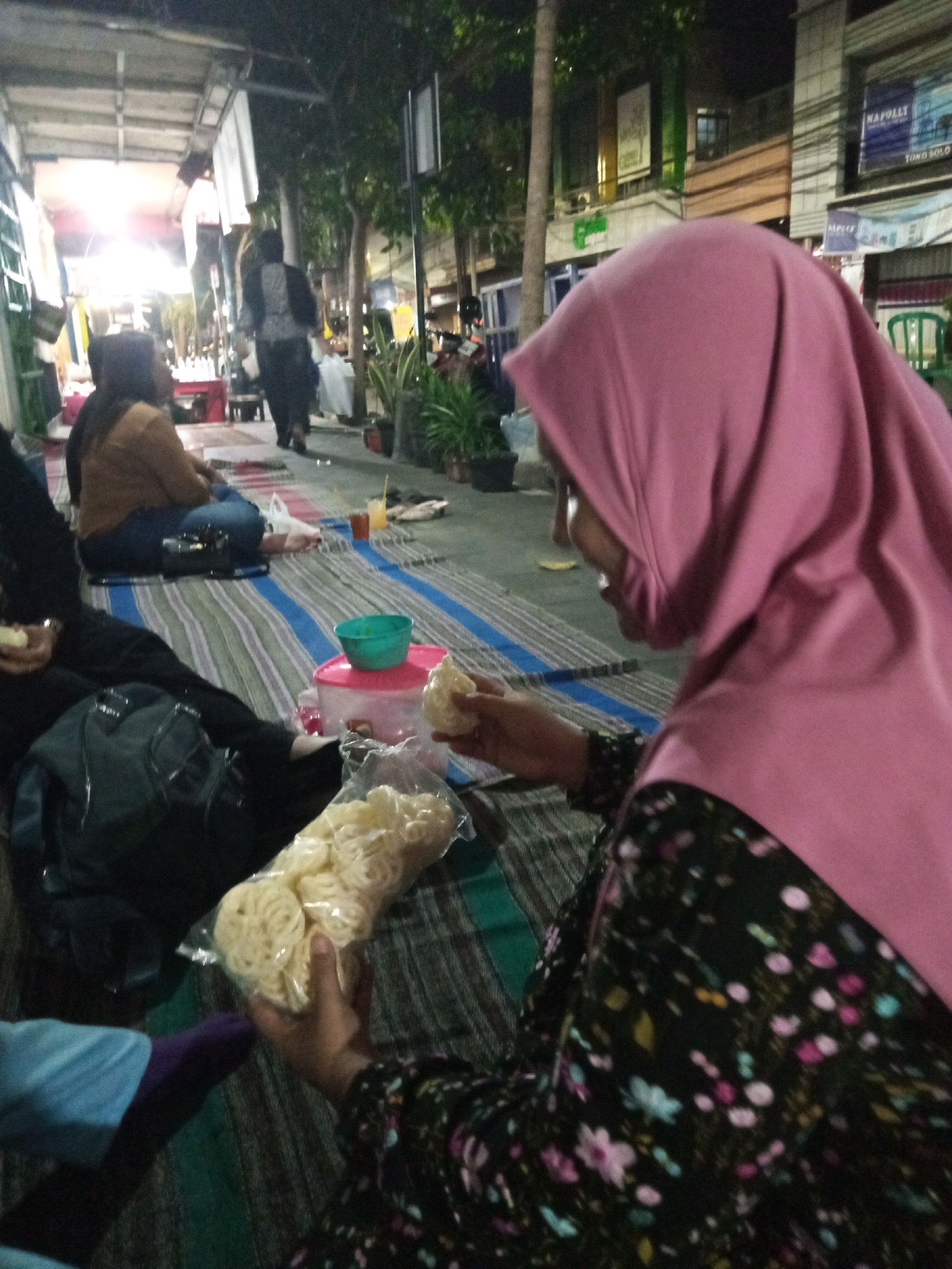 A group of people are gathered on a striped mat on the ground at night. The scene appears to be a street food stall or marketplace in Kediri City, Indonesia, with a covered walkway overhead and buildings visible in the background. The focus is on a woman in the foreground wearing a pink hijab and a floral patterned dress. She is holding a plastic bag filled with round, light-colored crackers and is in the process of eating one. To her left, another woman in a tan top and blue jeans is seated, facing away from the camera. A dark backpack is on the mat near her. To the left of the frame, a person in a blue and purple garment is also seated. A green bowl sits on a pink plastic container on the mat. The overall lighting is dim, suggesting it is nighttime, with artificial lights illuminating the area.