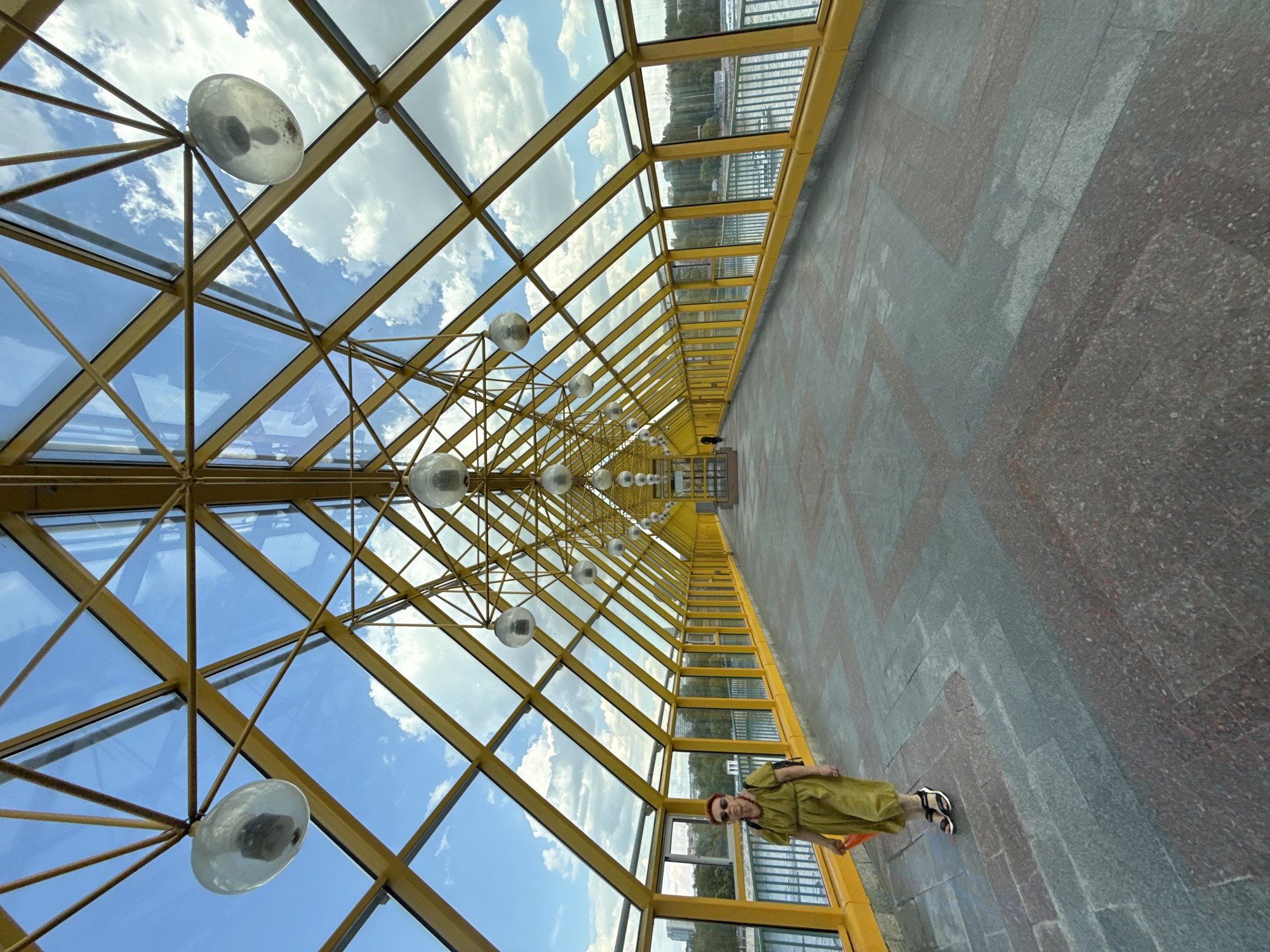The image captures the interior of a modern pedestrian bridge or covered walkway, characterized by a dominant yellow metal framework and extensive glass panels. The structure extends far into the distance, creating a strong sense of perspective and a tunnel-like effect. The ceiling, composed of triangular and rectangular glass panes, reveals a bright blue sky with scattered white clouds, indicating a clear, sunny day.
Along the center of the ceiling, a series of spherical, frosted glass light fixtures are suspended, receding in a line towards the far end of the walkway. The right side of the walkway (from the viewer's perspective) also features glass panels and yellow framing, offering a view of a body of water (likely a river) lined with trees and distant urban structures. The floor is covered with large stone tiles, arranged in a chevron or herringbone pattern using two contrasting shades of gray and reddish-gray.
A single person is visible on the right side of the walkway, standing relatively close to the foreground. This individual, appearing to be a woman with reddish-orange hair and sunglasses, is dressed in a loose-fitting, light olive green dress or tunic and black and white open-toed sandals. She holds an orange bag or object in her right hand and is positioned looking slightly away from the camera. There is no specific activity taking place beyond her standing and observing. No visible text is present in the image.