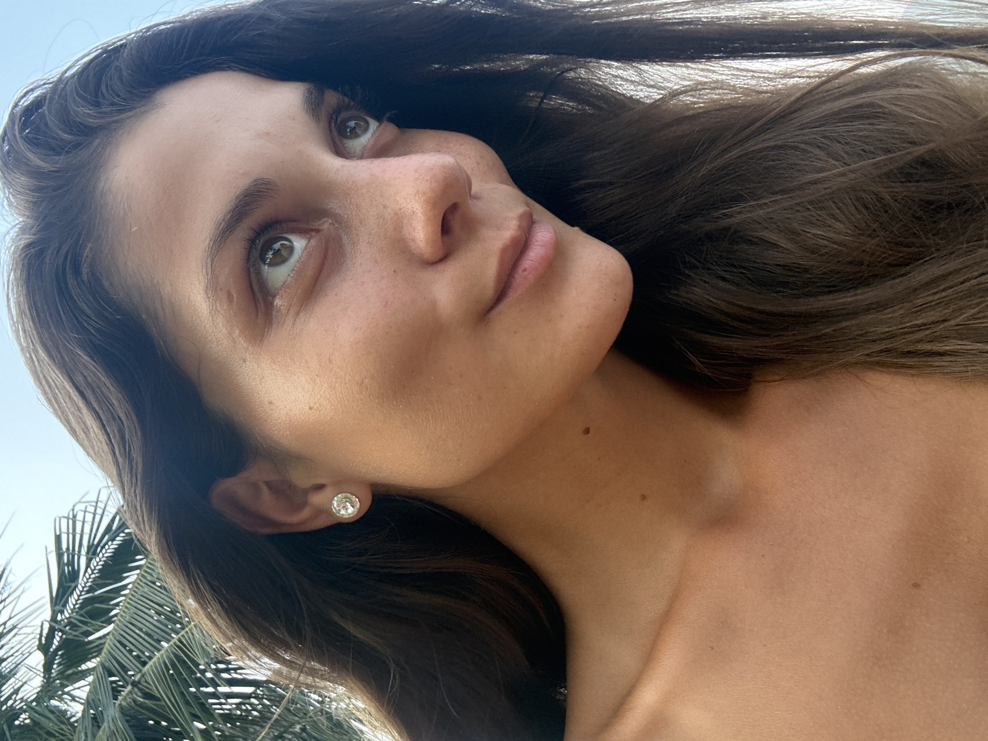 A woman with long brown hair and freckles gazes upwards in an outdoor setting. She is wearing a stud earring in her left ear. A palm tree is partially visible in the lower-left corner of the image. The location cannot be determined based on the image alone.