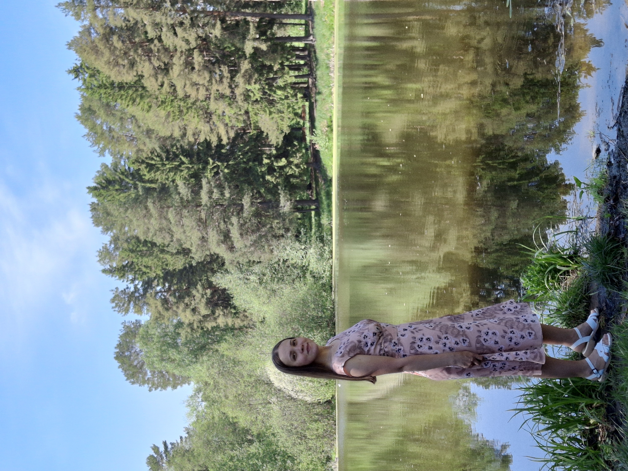 A full-length shot shows a woman in a light pink floral dress and white sandals lying on the grassy bank of a body of water. Her head is to the left, and her legs stretch toward the right. The body of water fills much of the upper half of the frame, reflecting the trees and sky. Trees line the opposite bank, and the sky is visible in the upper left corner. The image was taken in the city of Chaikovsky, Russia.