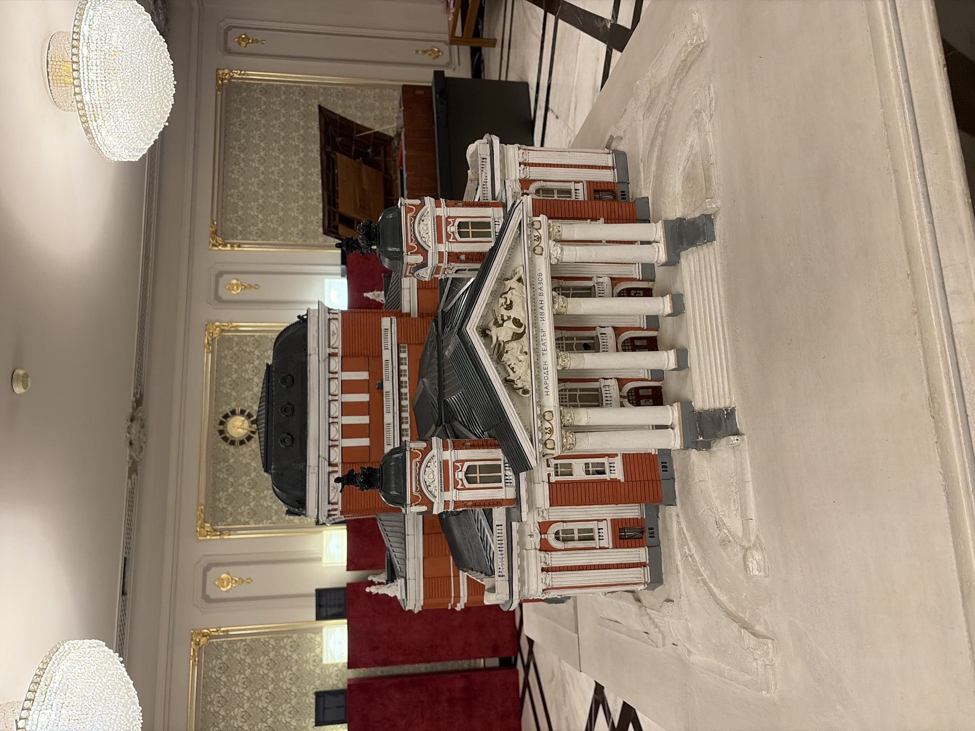 The image depicts a detailed architectural model of the Ivan Vazov National Theatre, a prominent landmark located in the heart of Sofia, Bulgaria. The model is displayed on a white platform, with the theatre itself rendered in its characteristic red brick and white column facade. The model accurately represents the theatre's classical architectural style, including its domed roof, portico with columns, and ornamental details.
In the background, the interior of a grand hall is visible, characterized by ornate gold trim on the walls, elaborate chandeliers hanging from the ceiling, and patterned wallpaper. Two large, round, crystal chandeliers are prominently featured, illuminating the space. Further in the background, a large, dark-colored object, possibly a grand piano or a piece of furniture, is partially visible, along with tall, red velvet panels that appear to be part of the room's decor or partition system. A clock is mounted on the wall in the upper background, a typical feature in such formal interiors. The floor of the hall is a patterned marble or tile. The overall impression is one of opulence and historical significance, befitting the representation of a national theatre. The model is the central focus, meticulously crafted to showcase the building's architectural grandeur.