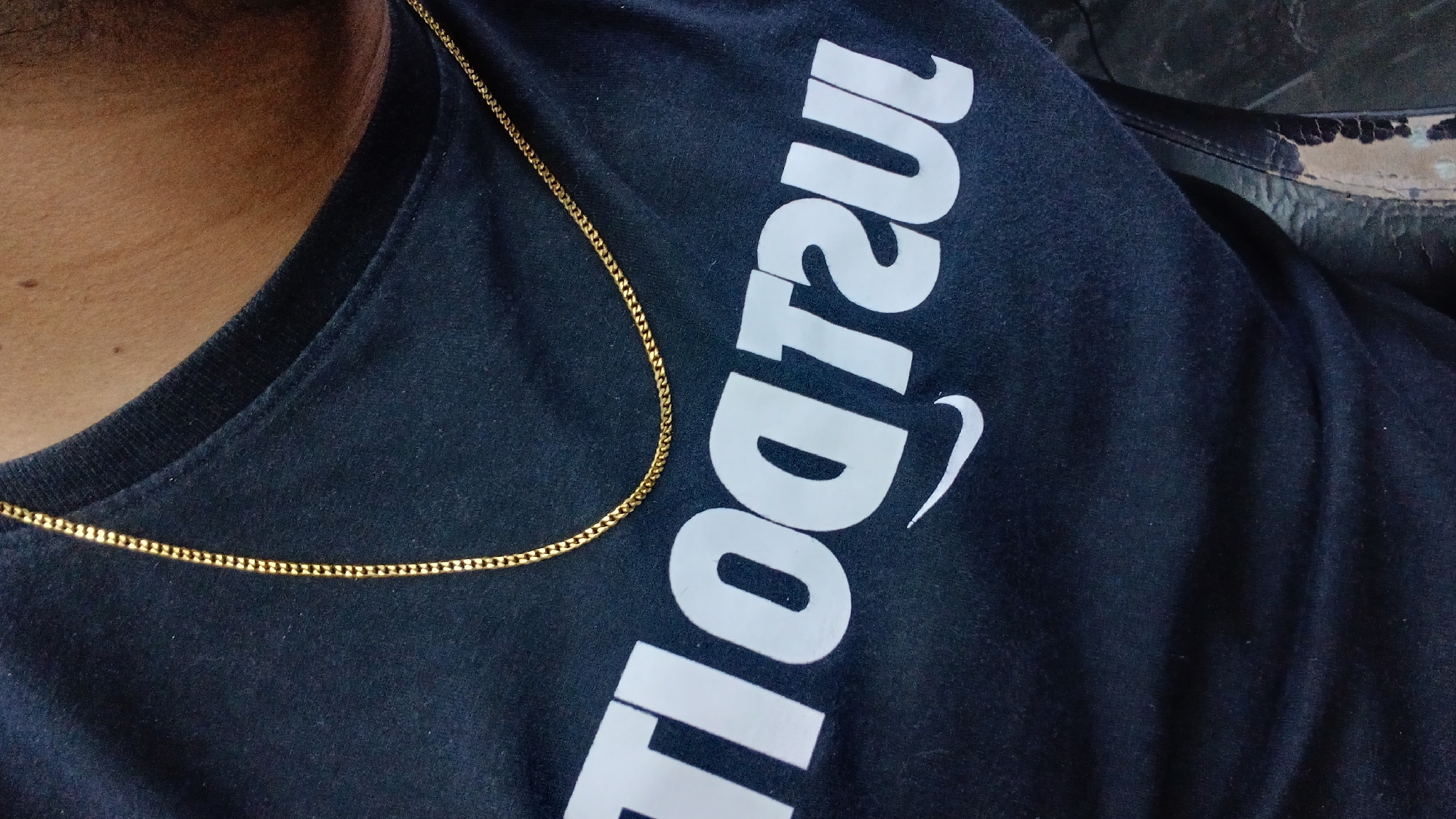A close-up shot shows a person wearing a dark blue t-shirt with white, large, rotated lettering that appears to be part of the Nike "Just Do It" slogan. A thin, gold chain rests around the person's neck. The person's skin is visible to the left of the collar. The background to the right contains what appears to be dark, textured material, possibly clothing. There is no indication of Juan Griego, Venezuela.