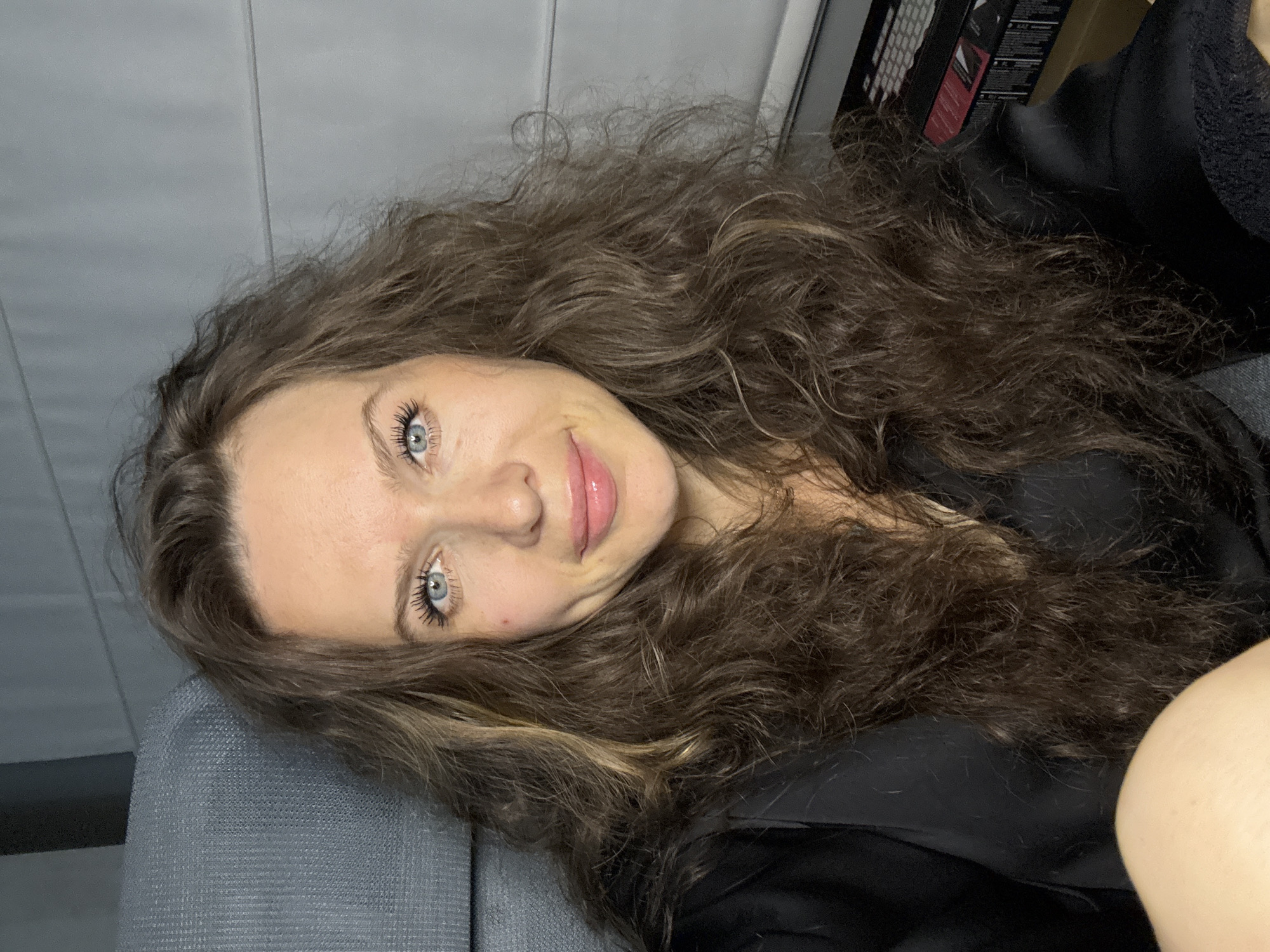 A woman with long, curly brown hair and blue eyes is lying down. She is wearing a black top and appears to be indoors, possibly on a couch. The background is a plain gray wall. The location is Unknown, Unknown.