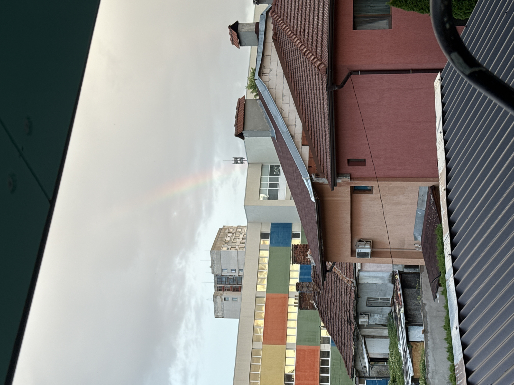 The image depicts a view of buildings and a rainbow against a cloudy sky, taken from an elevated position. In the foreground, a dark, corrugated metal roof is visible, partially obscuring the scene below. Behind this, a series of residential buildings are visible. The closest are two-story houses with terracotta tiled roofs. One house is painted a dark pink, with a dark brown downspout and a dark-paned window. Another adjacent house is a lighter, peachy color with white trim around its windows and an air conditioning unit mounted on the exterior wall. Further back, a more weathered, light blue structure is seen, also with an air conditioning unit, and what appears to be a shed or lean-to attached to it, with overgrown vegetation.
Dominating the middle ground is a long, multi-story building with a facade divided into colorful rectangular panels of yellow, orange, green, and blue, punctuated by rows of large windows. Above this, another, taller, grey building with visible windows and a somewhat aged appearance stands out. On top of this grey building, a telecommunications tower is visible.
In the sky, a faint rainbow is visible, arcing across the otherwise overcast and grey clouds. The overall impression is of a slightly overcast day in an urban or suburban setting, with the rainbow adding a touch of color and ephemeral beauty to the scene. The architectural styles suggest a location in Eastern Europe, possibly Bulgaria, given the common building materials and designs. The scene is devoid of any visible people. The viewpoint suggests the photo was taken from a balcony or a window of a higher building.