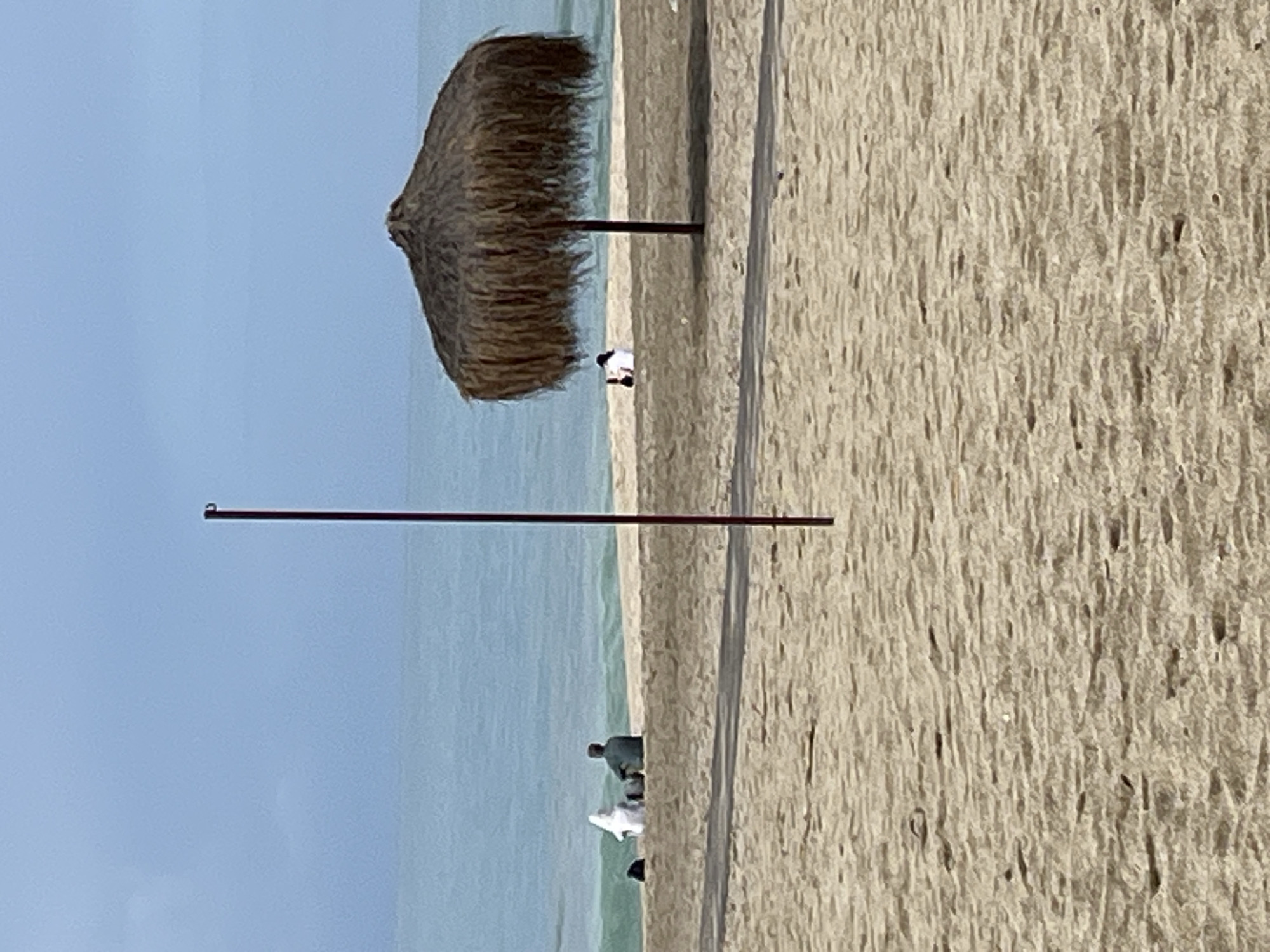 The image depicts a beach scene in Alanya, Turkey. A thatched-roof beach umbrella provides shade on the sandy beach. People are present near the shoreline, with one individual seated. The ocean stretches out to the horizon under a clear sky. Two metal poles stick up from the sandy ground in the foreground.
