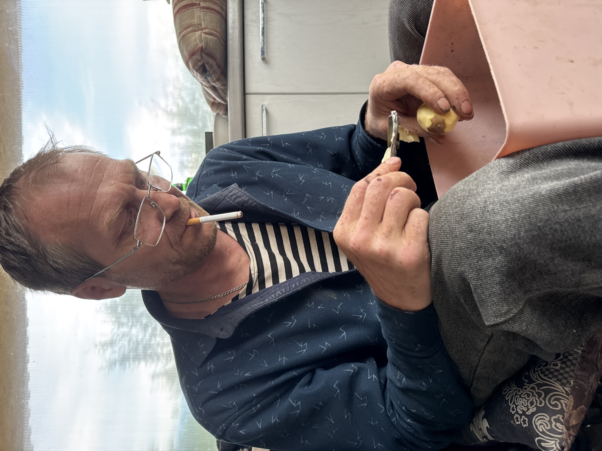In an indoor setting, a man with glasses and a cigarette in his mouth is peeling a potato with a vegetable peeler. The potato is held over a pink container. The man is wearing a striped shirt, a patterned navy blue jacket, and gray pants. In the background, there's a window and what appears to be a cabinet or cupboard. There is no definitive evidence in the image to establish the location in the city of Chaikovsky, Russia.