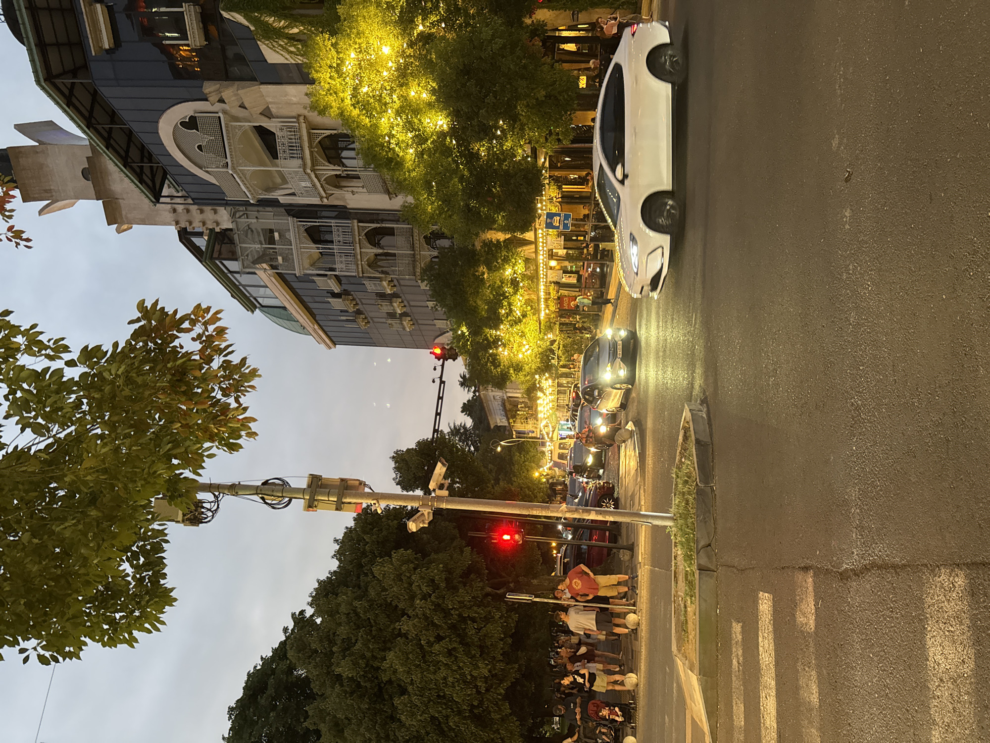The image depicts an evening scene at an intersection in Batumi, Georgia. A white sedan is driving on the right side of the road, and several other cars are visible in the background, suggesting moderate traffic. People are gathered on the sidewalk to the left, some with strollers, and others standing in groups. A traffic light shows red, indicating that vehicles are stopped. Trees line the street, and a building with ornate architectural details and illuminated lights is visible on the right side of the frame. The street is paved, and a pedestrian crossing is marked on the road. The sky is overcast, and the overall lighting suggests dusk or early evening.