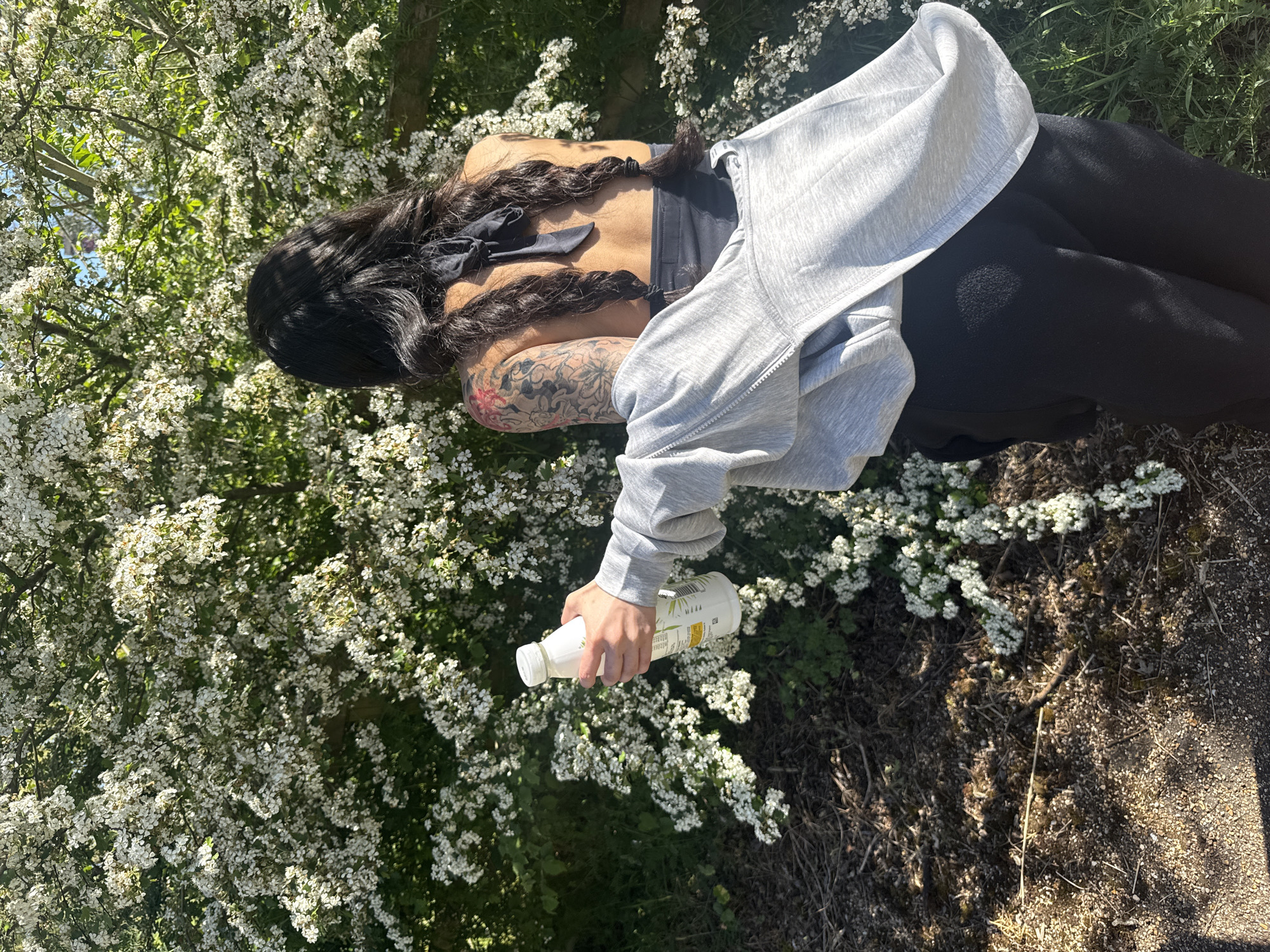 The image, rotated 90 degrees clockwise, presents a person viewed from behind, standing outdoors surrounded by nature. The individual has long, dark hair styled in two braids, each tied with a black ribbon. They are wearing a black sports-bra-like top, black trousers or leggings, and a grey zipped hoodie draped over their shoulders and left arm. An extensive tattoo, featuring floral or patterned designs with some red accents, is visible on their right arm.
The person is holding a white plastic bottle with a white cap in their right hand, extending it forward and slightly downwards. The bottle has a label with green and white graphics, but no specific text is legible.
The setting is an abundant bush or tree covered in numerous small white blossoms, suggesting late spring or early summer. Lush green foliage is visible both within and behind the flowering branches. The ground to the right and in the foreground consists of dark soil, dry leaves, and patches of gravel or a dirt path.
The scene is brightly lit by sunshine, indicating a clear day, likely morning or afternoon, with strong highlights on the flowers and hair, and visible shadows on the ground. The overall atmosphere is tranquil and natural. No specific London landmarks are visible, but the flowering vegetation is consistent with natural areas in the United Kingdom.
