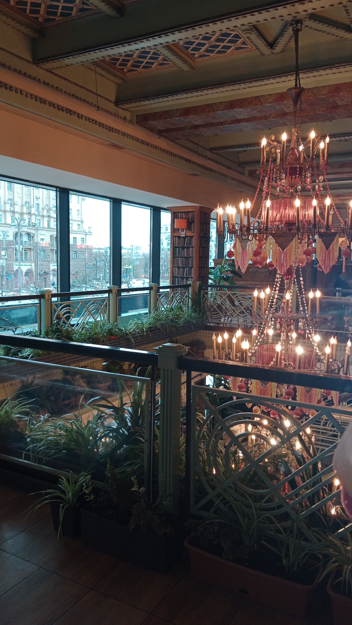 The image shows the interior of a multi-level space, possibly a restaurant or a public building, with a prominent chandelier hanging from a patterned ceiling. Large windows offer a view of a cityscape with buildings and a street. Indoor plants are arranged along the railings of the upper level, creating a sense of indoor greenery. A bookshelf is visible in the background, adding to the ambiance of the place. The architectural style of the building's interior suggests a classic or vintage design.