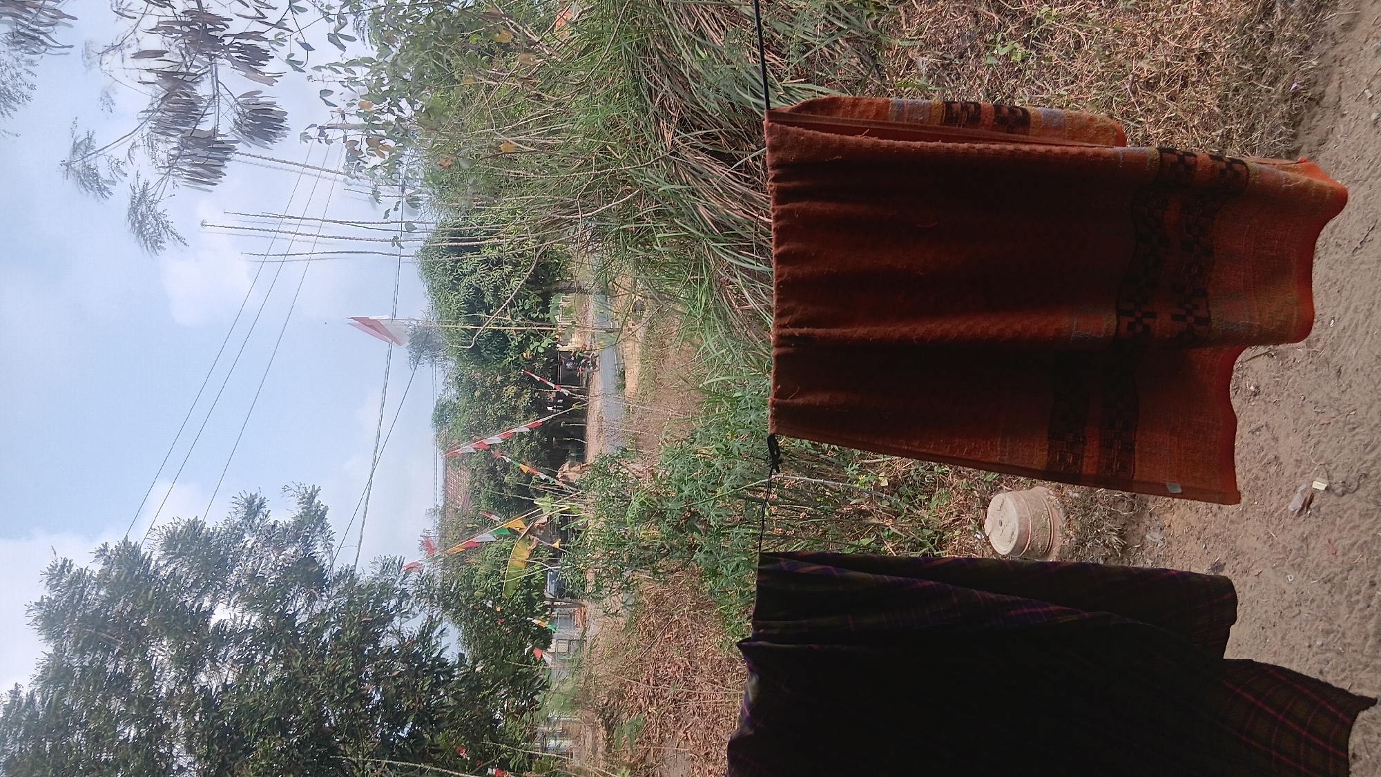 The image depicts a view of a rural roadside scene in Prabumulih, Indonesia, likely during daylight hours given the bright sky with scattered clouds. In the foreground, two pieces of fabric are hanging on a clothesline, obscuring the lower portion of the scene. The fabric on the left is dark with a plaid pattern, while the fabric on the right is a reddish-brown with a patterned border.
Beyond the clothesline, the environment transitions to dry, sparse vegetation and tall grasses. A road runs horizontally through the mid-ground, with a motorcycle visible traveling on it. On the other side of the road, there are houses with tiled roofs and numerous trees. Several Indonesian flags, red and white, are planted along the roadside and on poles, suggesting a celebration or national observance. The overall atmosphere is rustic and suggests a typical day in a semi-rural area.