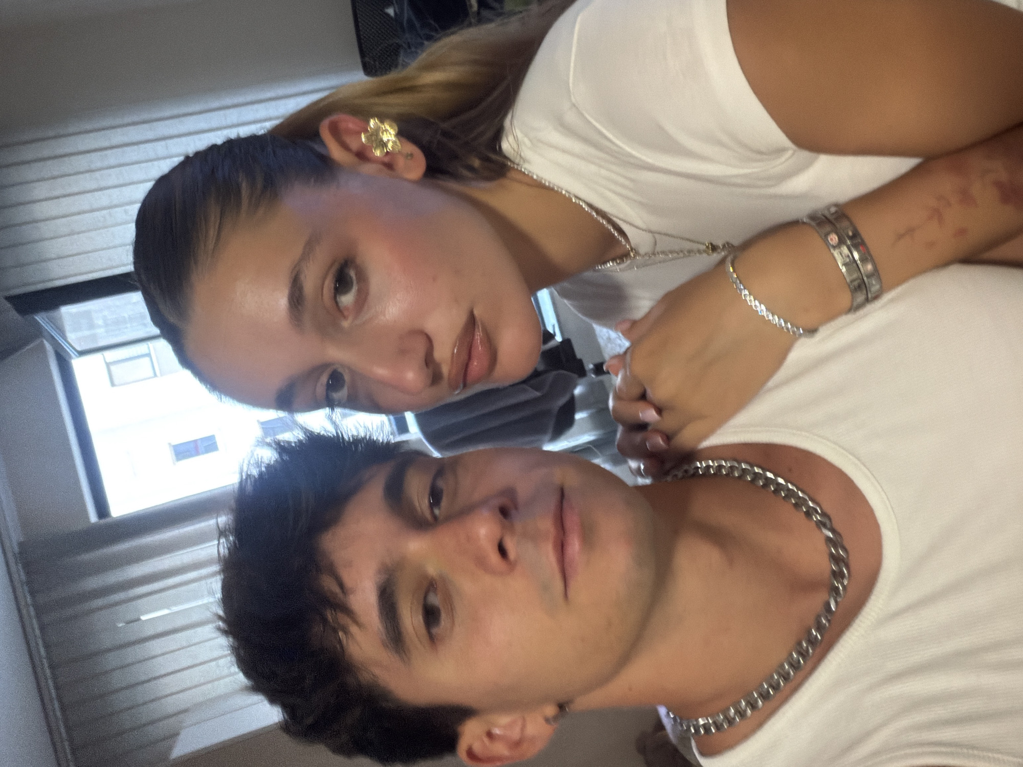 A close-up selfie of a young man and woman, both wearing white t-shirts, is captured in an indoor setting. The man, on the left, has dark, tousled hair and is looking directly at the camera with a neutral expression. He is wearing a thick silver chain necklace. The woman, on the right, has her hair pulled back in a ponytail, revealing a large, gold, flower-shaped earring in her right ear. She is wearing a pearl necklace and a diamond bracelet, and her left hand rests on the man's shoulder. The background consists of vertical blinds, a window with a view of a building, and some indistinct objects on the right. The lighting suggests it is daytime. The location is specified as Sofia, Bulgaria, though there are no visual cues in the image to confirm this.
